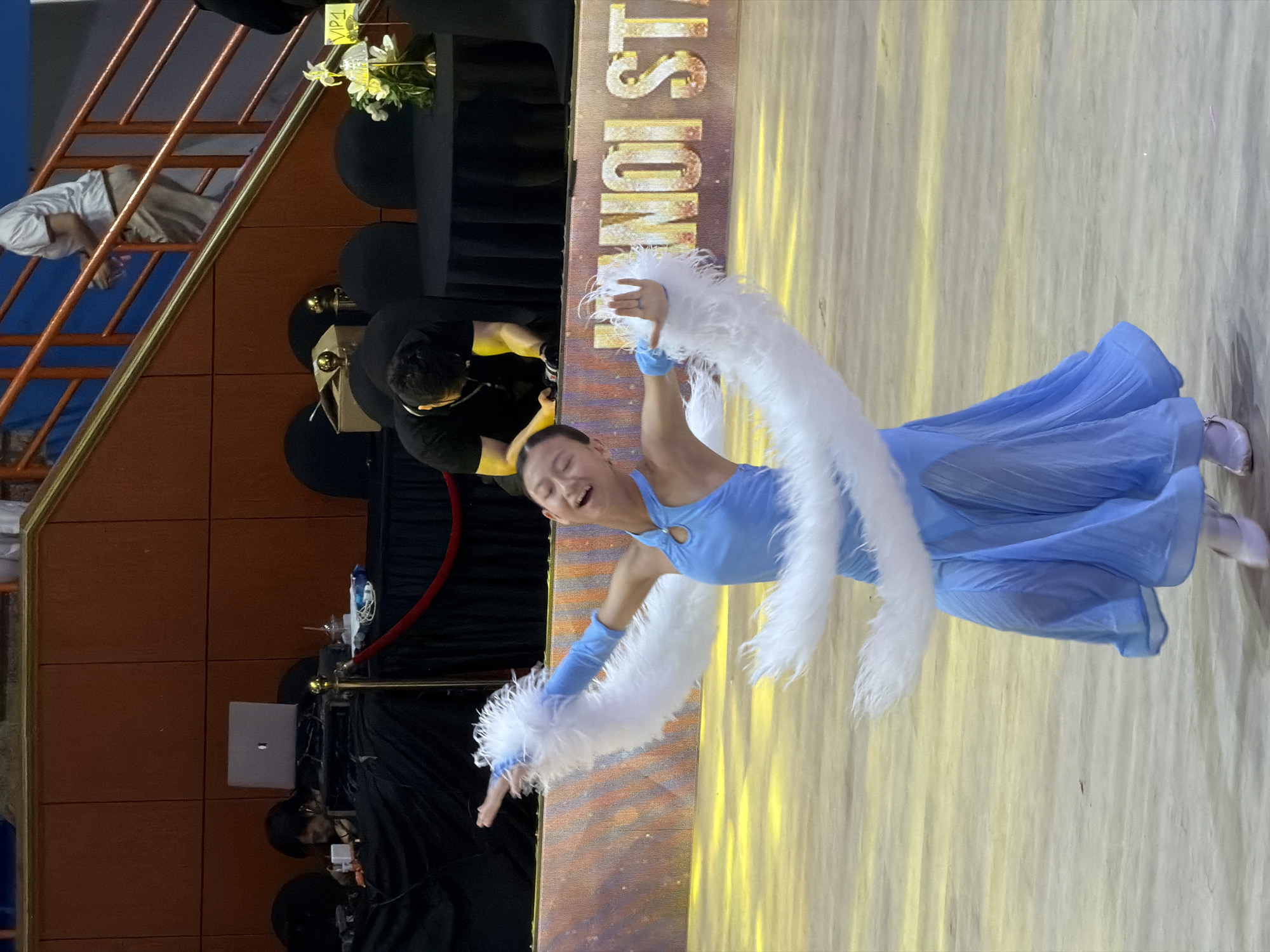 In Hanoi, Vietnam, a young female dancer in a light blue dress and white feathered arm coverings performs on a wooden floor with yellow lighting. She is mid-leap with her mouth open in a shout or laugh. Behind her, a banner with "HANOI STA" visible is illuminated. In the background, several individuals are seated at round tables draped in black cloth. One person, wearing a black shirt, is crouched down, looking at something on a table. Further back, another person in a white t-shirt walks up a set of orange stairs. There is also a laptop visible on a table to the left.