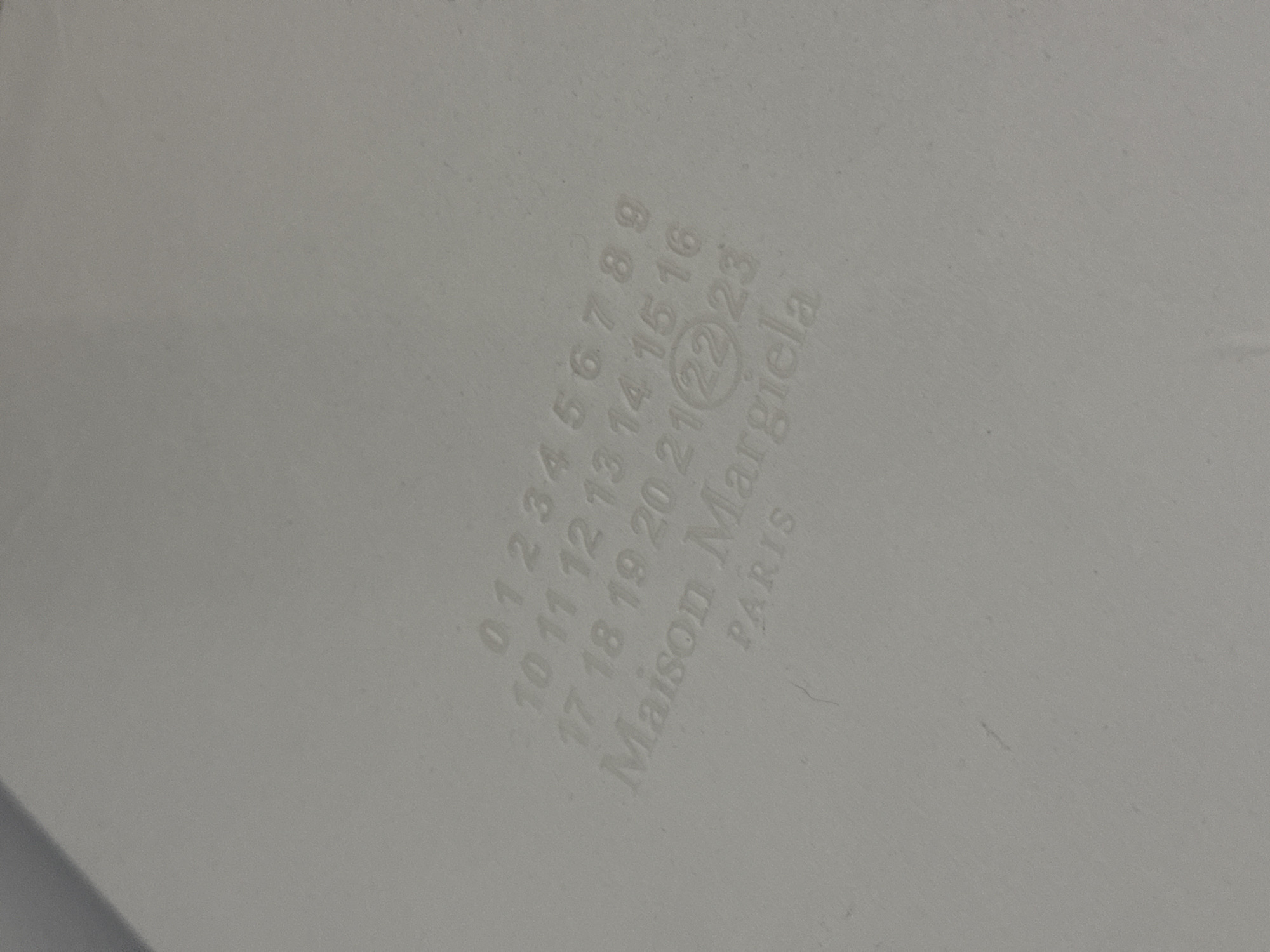 The image shows a close-up of a white surface with the Maison Margiela logo printed on it. The logo includes the numbers 0 to 23 arranged in a grid, with the number 22 circled. Below the numbers, the words "Maison Margiela" and "PARIS" are printed. The image is slightly out of focus and the lighting is somewhat diffused. There is no information about the location in the city of Burgas, Bulgaria, or any other details about people, subjects, objects, actions, interactions, or the environment.