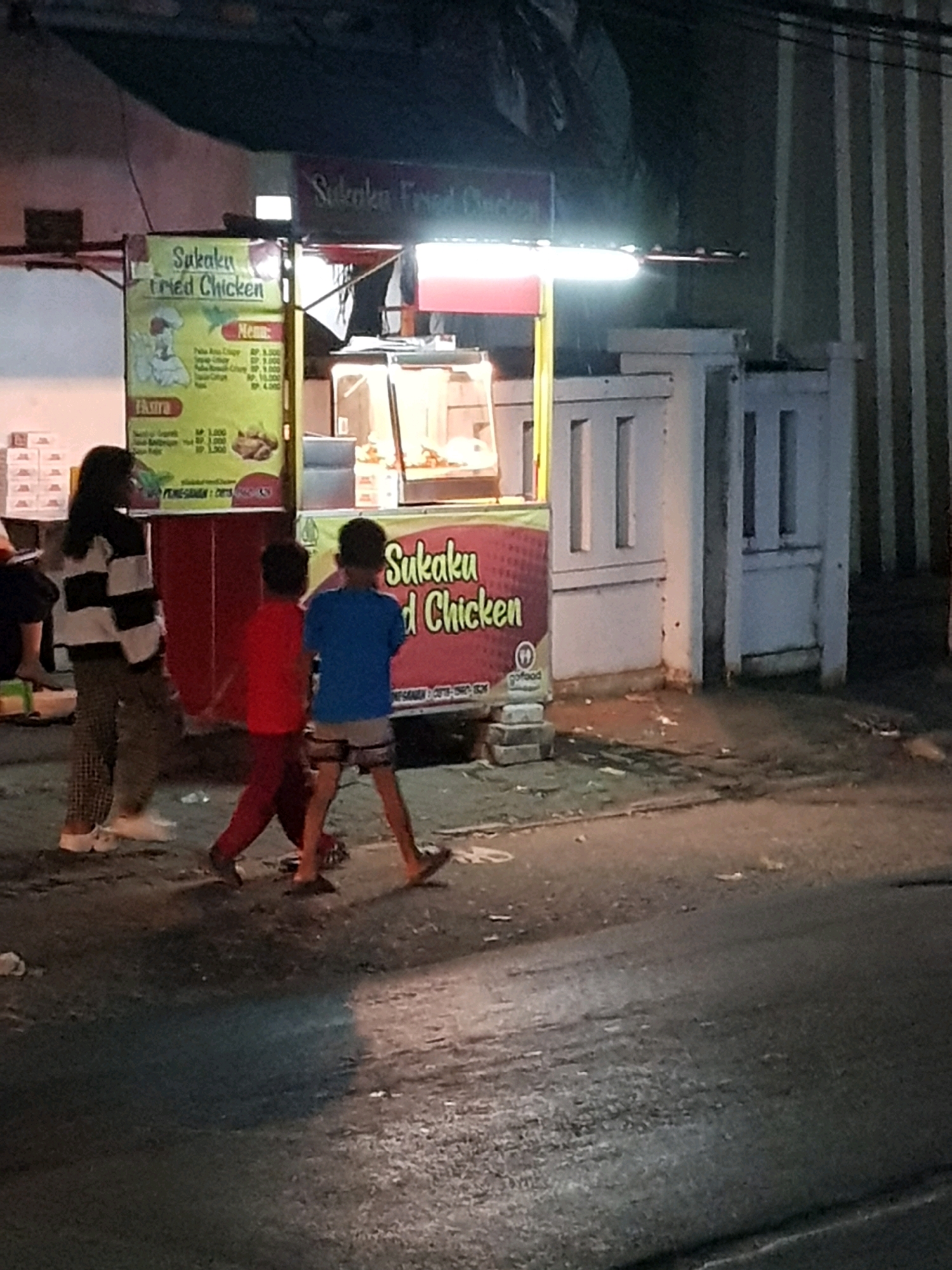 The image captures a nighttime scene at a "Sukaku Fried Chicken" food stall in Setiamekar, Indonesia. The stall features a lit menu board displaying prices for chicken and side dishes, as well as a display case with food. In front of the stall, three individuals are visible. A woman wearing a striped long-sleeved shirt and checkered pants stands on the left, facing away from the camera. To her right, two young boys walk away from the stall. One boy wears a red shirt and dark pants, while the other wears a blue t-shirt and shorts. The ground in front of the stall appears wet and somewhat dirty, with debris scattered on it. The surrounding environment suggests an outdoor urban setting, possibly a street or roadside, with a building structure visible behind the food stall.