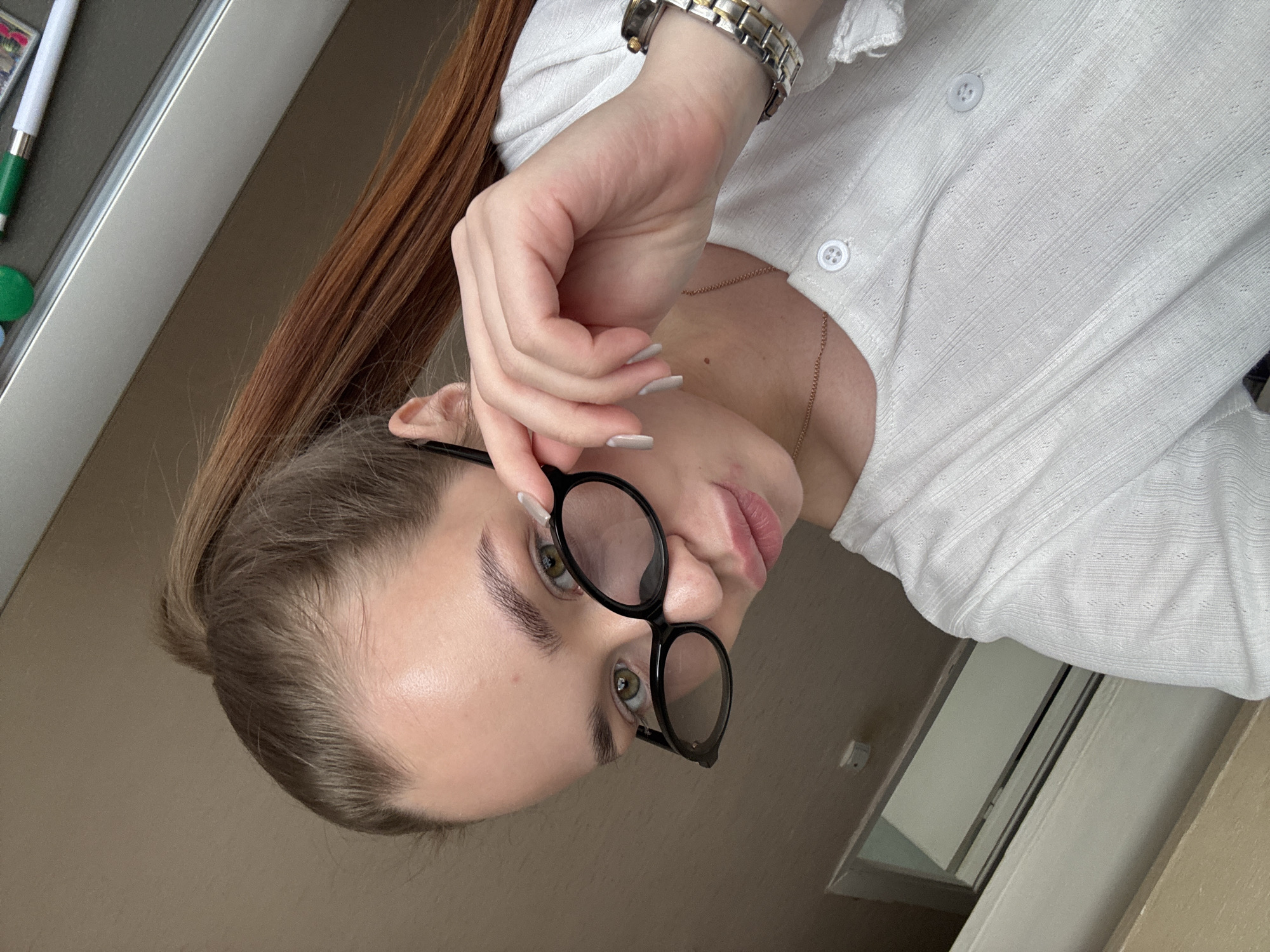 A young woman is depicted in an indoor setting. She has fair skin, brown hair pulled back into a ponytail, and is wearing black, round glasses. Her nails are manicured with a light color. She is wearing a white, button-down shirt and a watch. The background suggests an interior space, with a partial view of a window. There is no explicit information to confirm the exact location in the city of Chaikovsky, Russia.