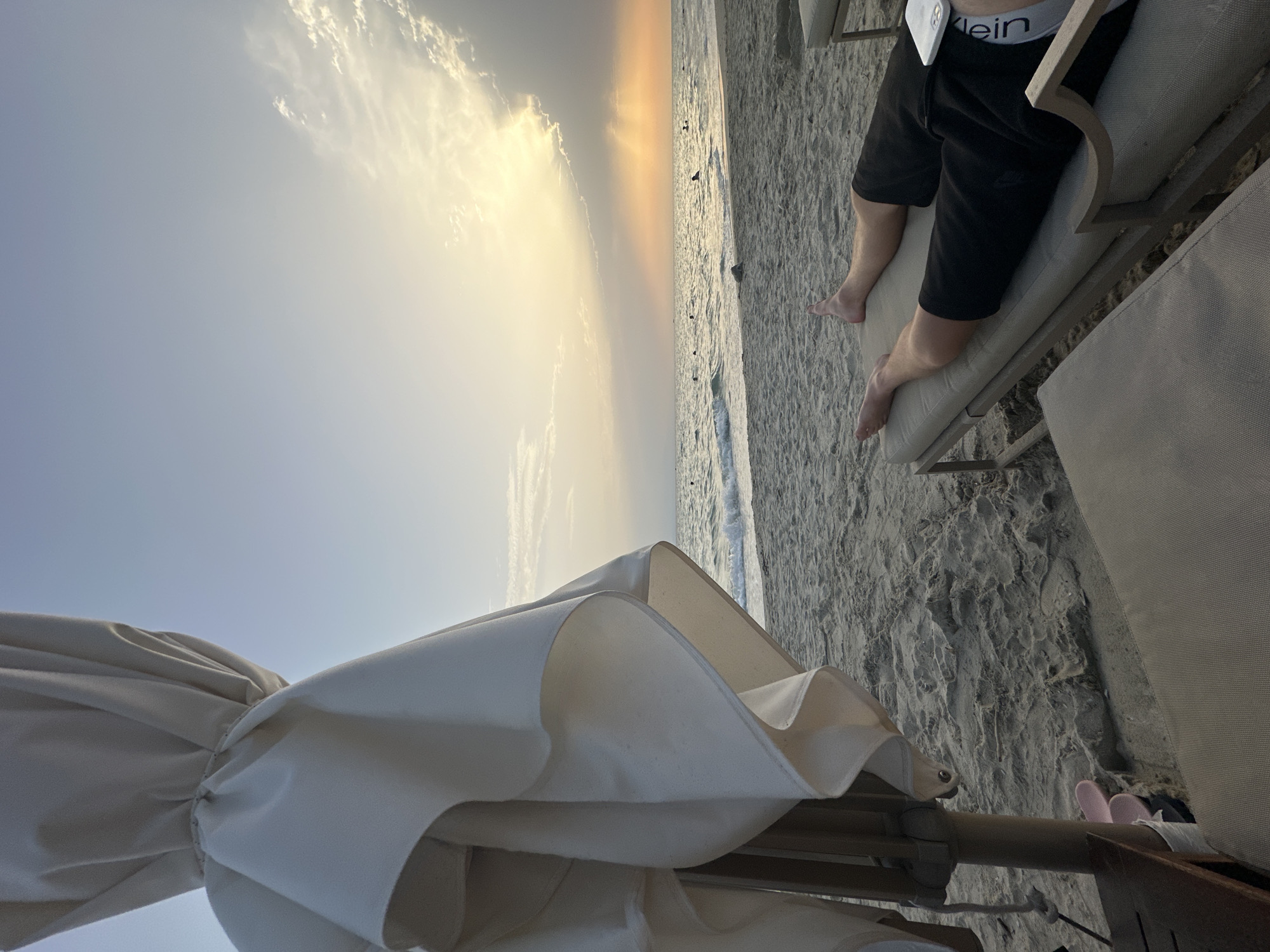 The image depicts a serene beach scene at sunset. In the foreground, a person is lying on a lounge chair, their legs extended, wearing black shorts. A closed umbrella is partially visible on the left, providing shade. The sandy beach stretches towards the ocean, where gentle waves are rolling in. Several small figures can be seen in the water, some appearing to be swimming. The sky is painted with warm hues of orange and yellow as the sun descends, casting rays of light through the clouds. The overall impression is one of relaxation and tranquility on a Grecian beach. The location is likely a resort or public beach within Kassandra Municipal Unit, Greece, given the presence of lounge chairs and umbrellas typical of tourist areas.