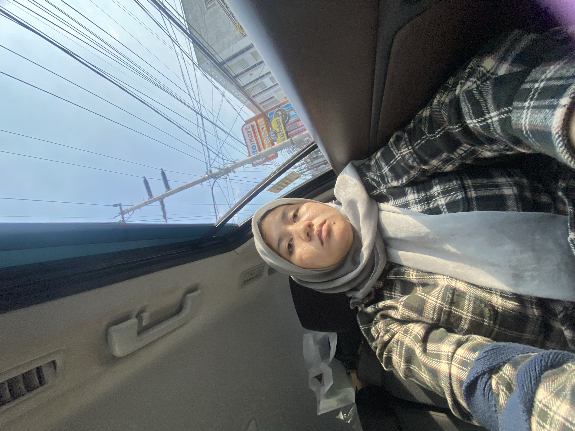 A woman wearing a grey hijab and a plaid shirt is sitting in the back seat of a car. The car's interior is visible, including a handle on the ceiling. Outside the car window, there are power lines, a utility pole, buildings, and a sign for "Alfamart". The sky is clear and blue. The woman is looking at the camera with a neutral expression. The image appears to have been taken during the day, likely in a populated area of Banjarmasin, Indonesia, given the presence of the Alfamart sign.