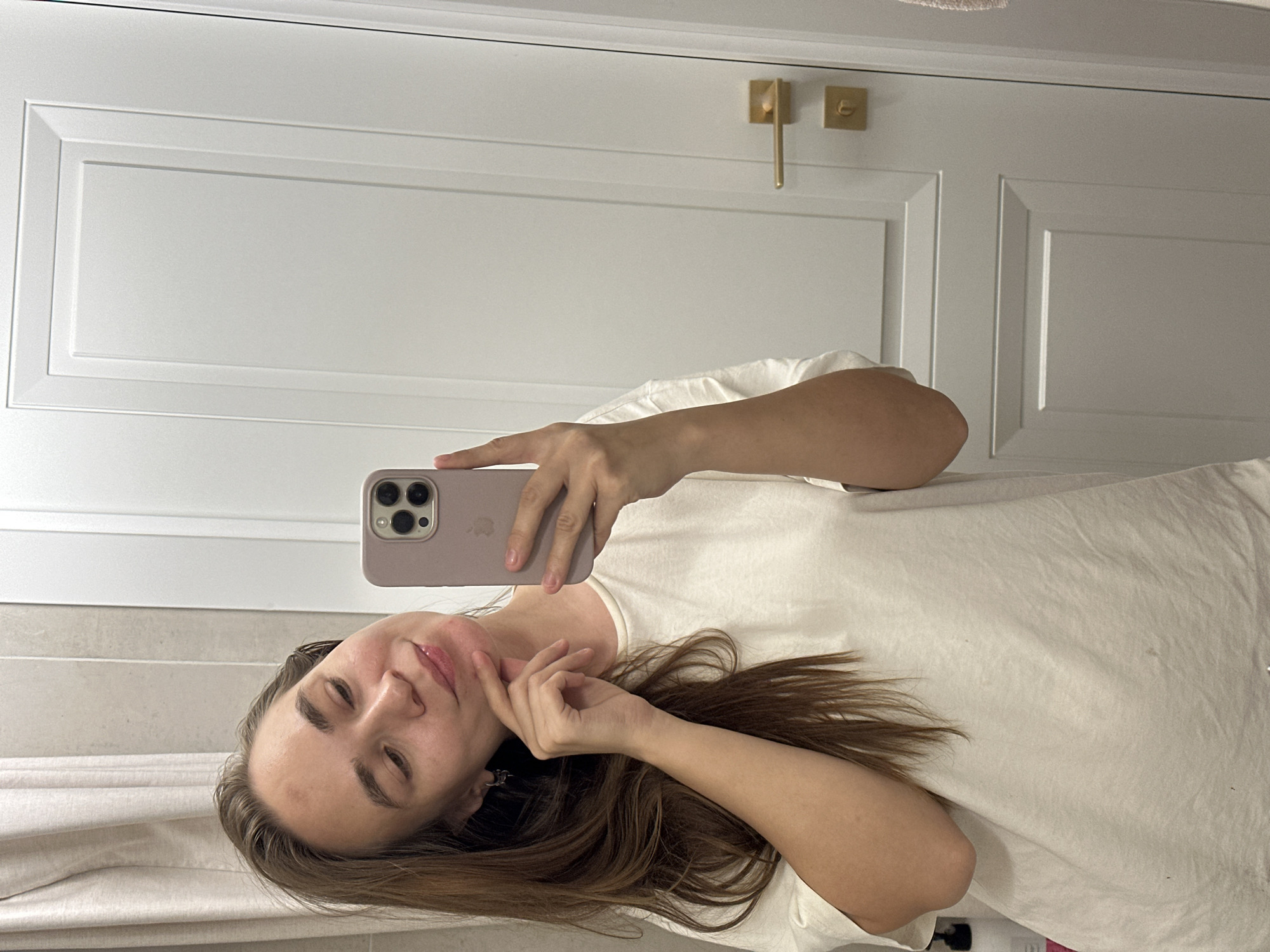 A young woman is taking a selfie in what appears to be a bathroom. She is holding a lavender iPhone in her right hand and is looking at the camera. She is wearing a white t-shirt. Her long brown hair is draped over her shoulder. Behind her is a white door with gold hardware. The presence of a door and presumed bathroom is assumed, not explicitly proven. There is no evidence to suggest that the location of the image is in Kharnavy, Russia.