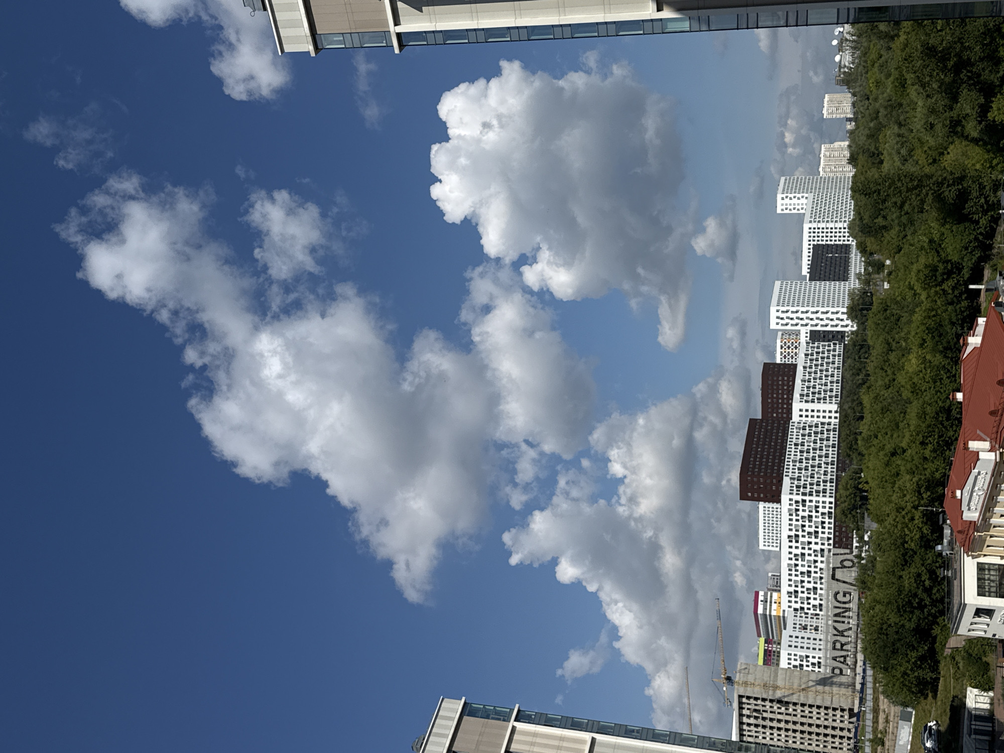 The image captures a panoramic view of a modern urban landscape under a bright blue sky dotted with fluffy white clouds. The scene features several tall residential buildings with contemporary architecture, characterized by clean lines, large windows, and a mix of white, brown, and grey facades. One prominent building in the mid-ground displays the word "PARKING" in large, dark lettering against a lighter background. Construction cranes are visible, suggesting ongoing development in the area.
A dense green forest lines the foreground and mid-ground, providing a natural contrast to the urban structures. In the lower portion of the image, rooftops of smaller buildings with red and white finishes are visible, hinting at a transition from newer constructions to older ones or residential areas. A few parked cars are present near the edge of the forest on the left side of the frame.
The overall impression is one of a dynamic and developing city, possibly located in Moscow, Russia, given the architectural style of the buildings and the presence of green spaces within the urban environment. The weather appears to be pleasant and sunny. There are no visible people in the frame.
