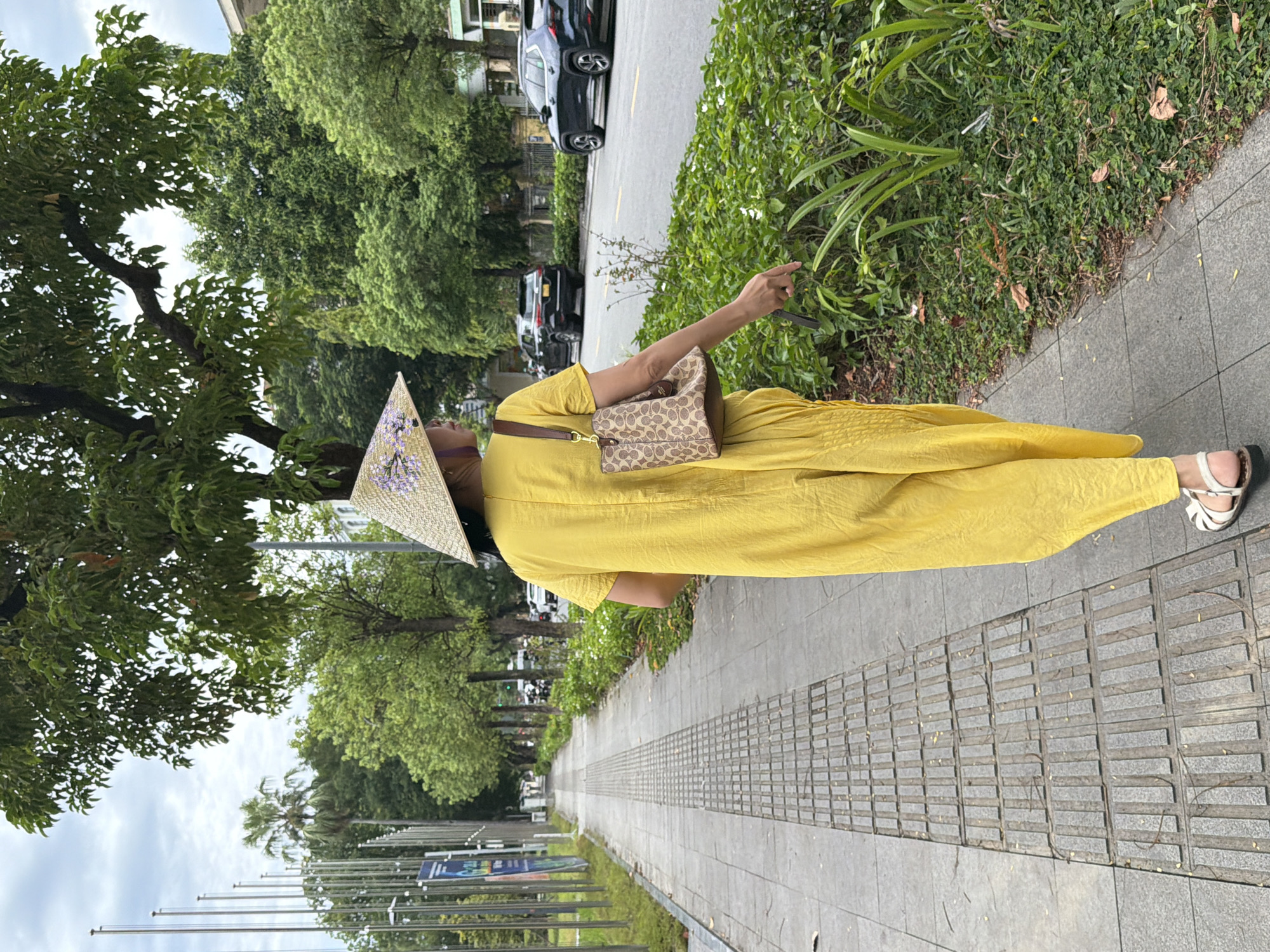 A person wearing a yellow jumpsuit and a conical hat walks along a paved sidewalk next to a street in Thuận Hóa Ward, Vietnam. The person is carrying a brown patterned handbag and is seen from behind, walking away from the camera. The sidewalk has textured tactile paving for the visually impaired. The street has several cars and is lined with trees and green foliage. In the background, tall poles with flags are visible. The sky is overcast.