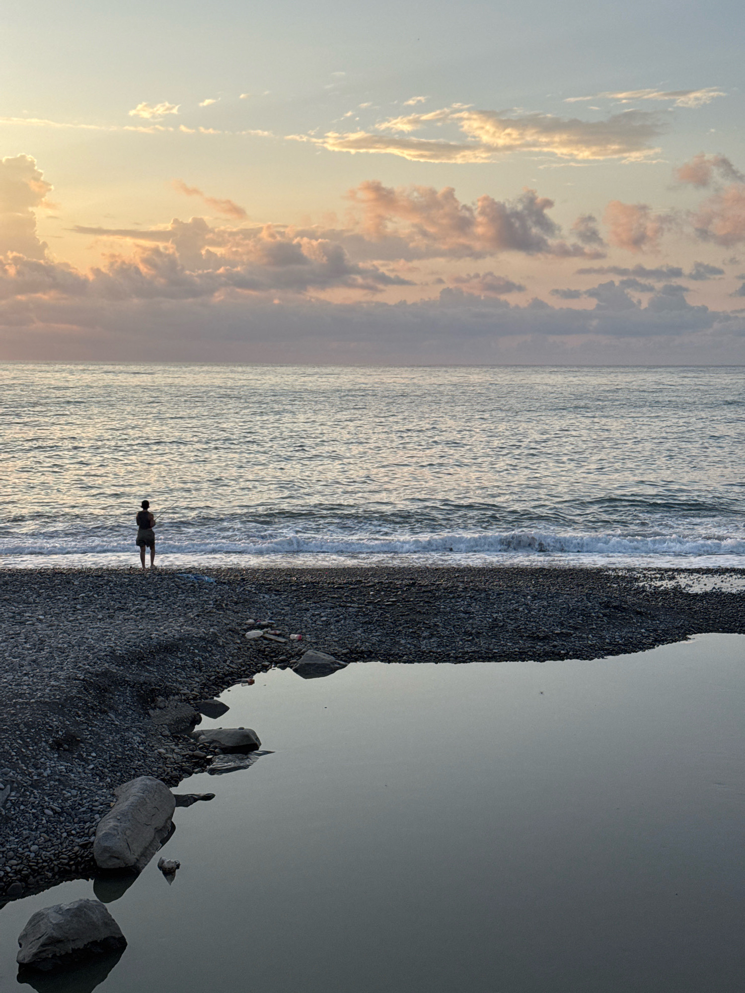 A lone figure stands on a pebble beach in Batumi, Georgia, facing the Black Sea at sunset. The sky is a canvas of soft oranges, pinks, and blues, with scattered clouds catching the last rays of the sun. Gentle waves lap at the shore, creating a foamy white line. In the foreground, a still pool of water reflects the muted colors of the sky and the surrounding stones. A few pieces of debris are visible on the beach, including plastic bottles. The figure, silhouetted against the sea, appears to be fishing. The scene conveys a sense of tranquility and solitude.