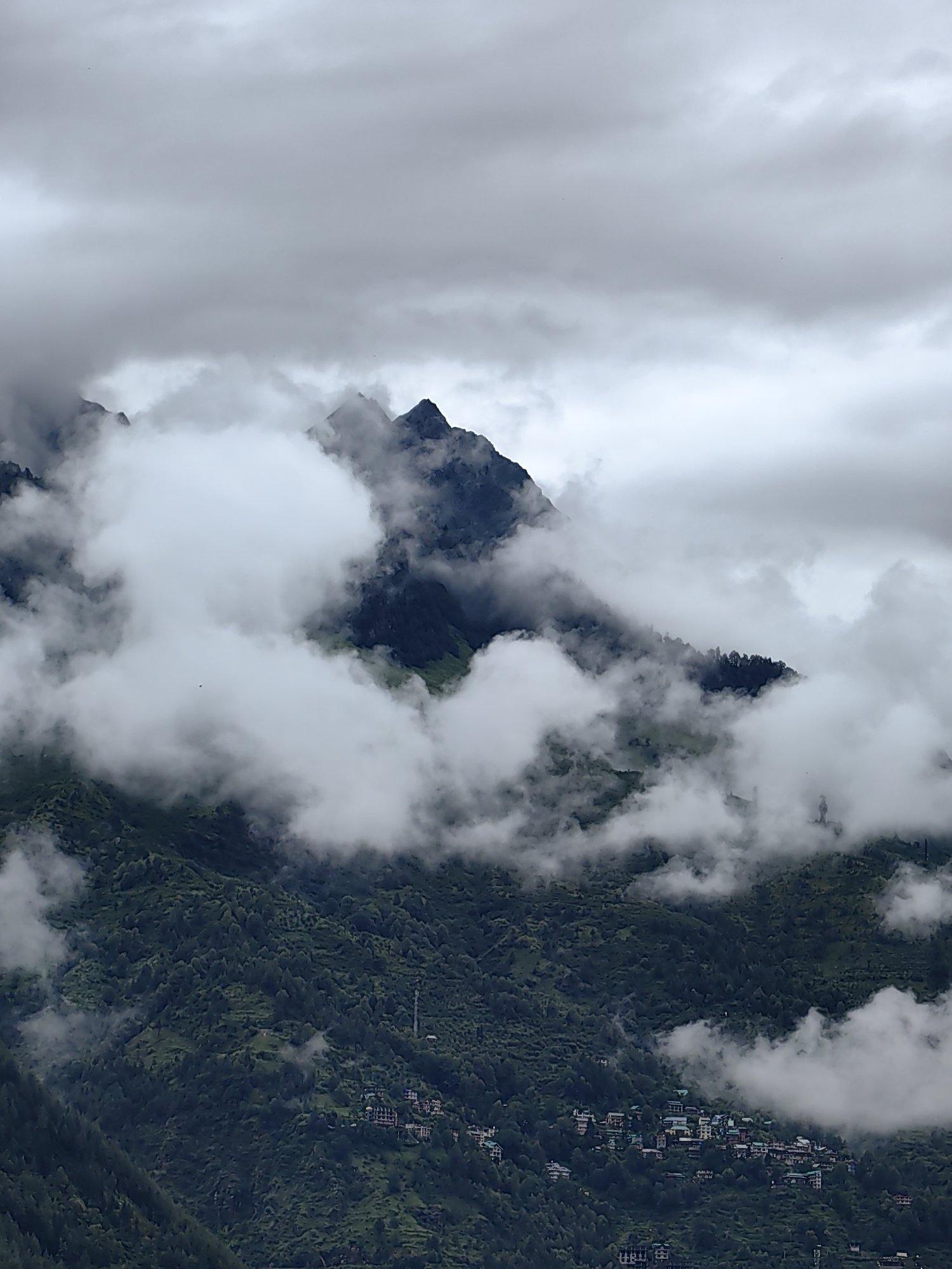 The image depicts a misty mountainous landscape in Vashisht, India. The foreground is dominated by dense, dark green vegetation covering steep slopes. Small clusters of buildings, likely homes and guesthouses, are visible nestled into the hillsides. These structures are characterized by multi-story designs with colorful roofs. A cell tower is also present on one of the slopes.
In the midground and background, towering mountains rise, their peaks shrouded in thick, swirling clouds. The clouds create a dramatic and ethereal atmosphere, partially obscuring the rugged mountain terrain and lending a sense of depth to the scene. The sky above is overcast with more layers of clouds, contributing to the overall moody and diffused lighting. There are no visible people or animals in the image.