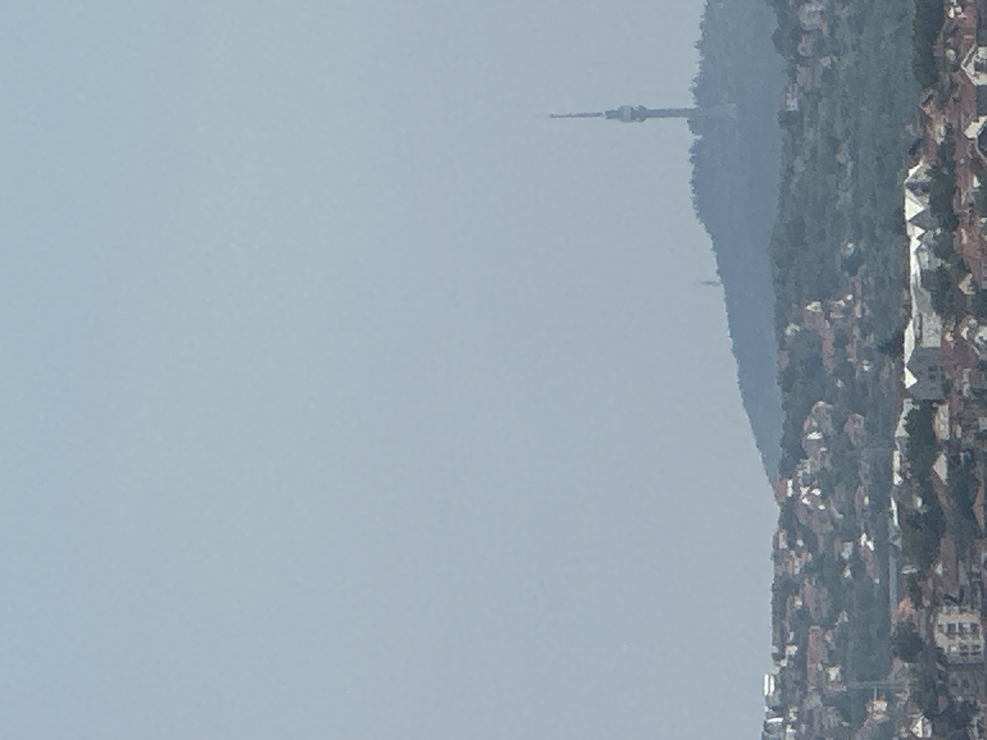 This media file depicts a hazy, possibly overcast day in Belgrade, Serbia. The scene is dominated by a vast expanse of pale blue sky, suggesting either early morning or late afternoon light, or potentially a significant amount of atmospheric haze.
In the distance, a slender, tower-like structure is visible, its top piercing the sky. This could be a telecommunications tower or a monument. It stands against a backdrop of a densely wooded hill that slopes down towards a city.
The city itself appears to be built on the hillside, with a cluster of buildings visible. The architecture suggests a European setting, with rooftops and facades rendered in muted tones, likely due to the distance and the atmospheric conditions. Some of the buildings appear to be residential, while others may be commercial. The overall impression is one of a sprawling urban area nestled into the natural landscape.
The orientation of the image is rotated, with the horizon appearing vertically. The weather seems to be calm, with no indication of strong winds or precipitation. The hazy conditions contribute to a soft, diffused light across the entire scene. There is no visible text in the image.
