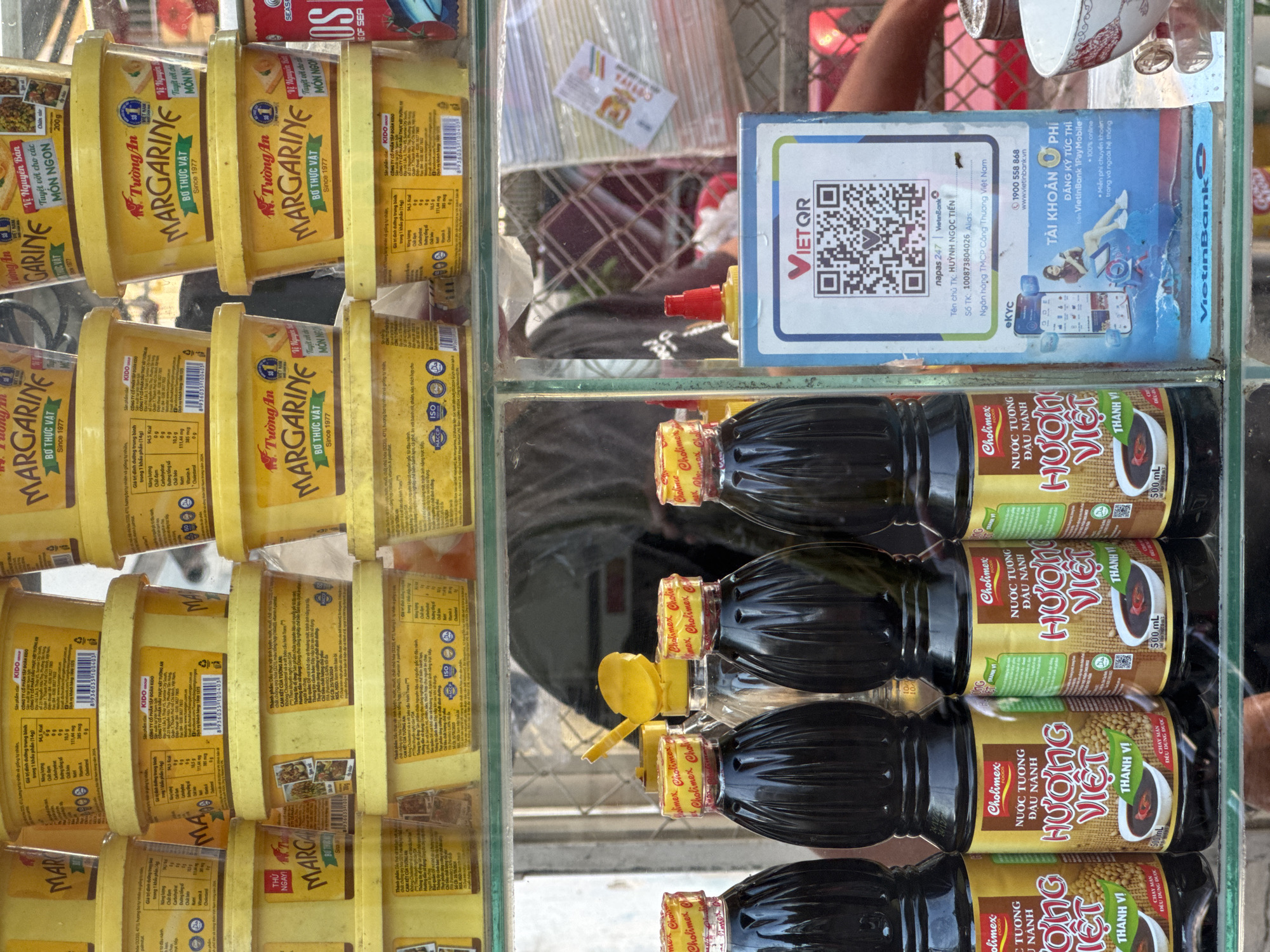 The image depicts a street vendor's stall in Nha Trang Ward, Vietnam, showcasing a variety of food products and merchandise. In the foreground, several bottles of Chollmex Huong Viet soy sauce are neatly arranged. The bottles are dark brown with yellow labels featuring the brand name "Chollmex," the product name "NUOC TUONG HUONG VIET," and "THANH VI" indicating a savory flavor. Some bottles have red caps and others have yellow caps. Behind the soy sauce bottles, a clear plastic bottle with a yellow cap is visible.
The middle ground of the image is dominated by stacks of yellow tubs of margarine. These tubs are labeled with Vietnamese text and feature branding that includes "MARGARINE," "BO THUC VAT" (vegetable butter), and "VI NGUYEN BAN" (original flavor). Some tubs also display nutritional information and certifications like HACCP and ISO.
In the background, partially obscured, are more products and some general store items. A red can with "OS" and "SEA" is visible on the far right. A portion of a QR code for "VIETQR" and information about VietinBank, including account details and a phone number, are displayed on a blue and white stand. This suggests that the vendor accepts digital payments.
The overall environment appears to be an outdoor stall, likely on a street, with the goods displayed on shelves. The lighting suggests it is daytime. There are no people clearly visible interacting with the products, but a blurred figure is visible behind the margarine tubs, likely the vendor.
