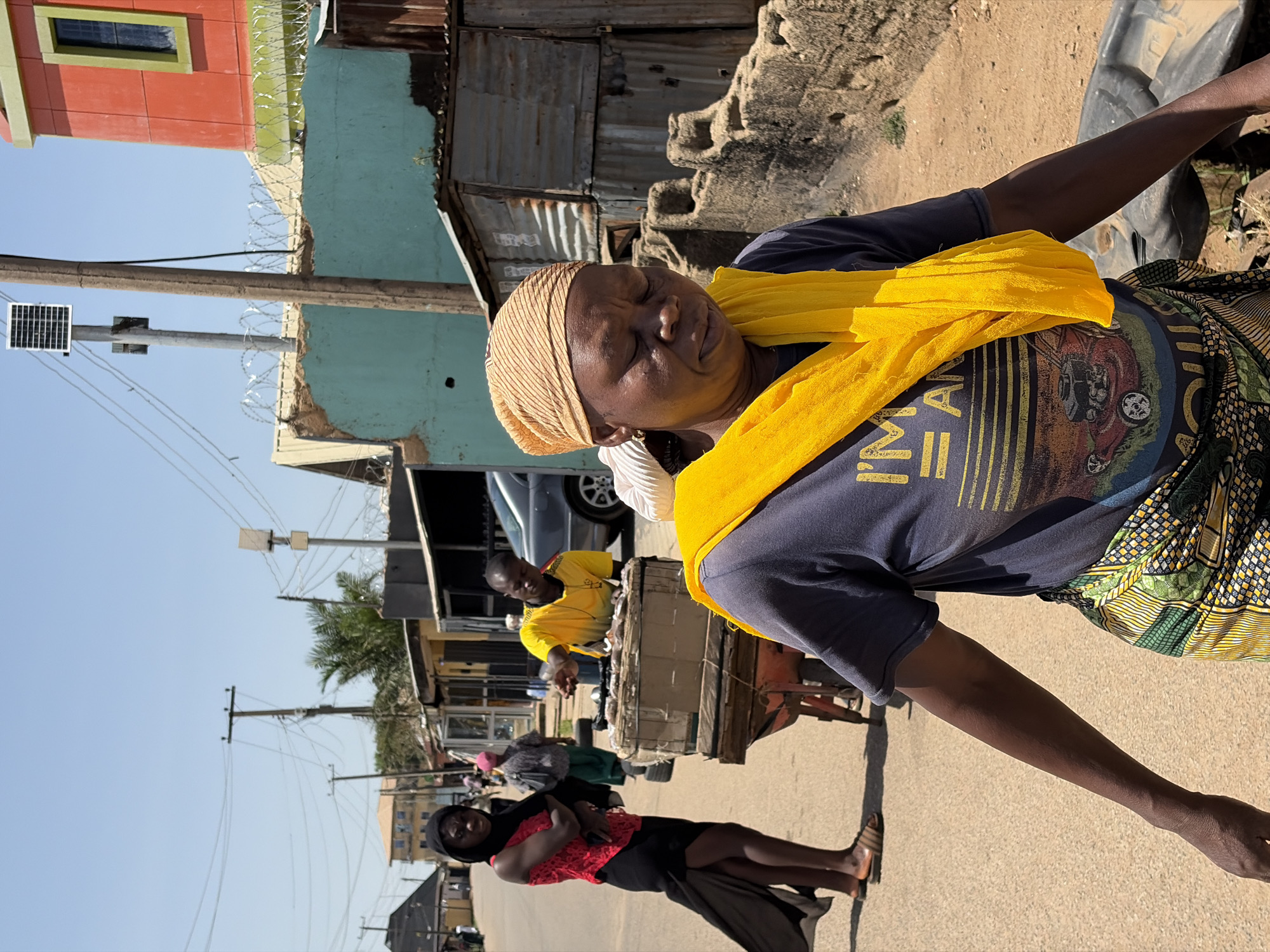 This image captures a vibrant street scene in Jos, Nigeria, under a bright, clear sky, likely mid-day due to the strong sunlight. The main subject is a dark-skinned woman, prominently featured in the foreground, looking downwards with her eyes closed in what appears to be a moment of rest or contemplation. She wears a light brown headwrap with thin stripes, a bright yellow scarf draped over her shoulders, and a dark blue t-shirt. The t-shirt bears a graphic design featuring a vintage-style car, possibly a Volkswagen Beetle, with partial text reading "I'M A". A colorful, patterned fabric, predominantly green, yellow, and black, is wrapped around her lower body.
In the mid-ground, behind the main woman, another person in a yellow shirt is visible, appearing to stand behind a wooden cart or market stall. Further down the street, a woman in a black head covering and a red top stands by the roadside. The street is lined with various buildings showcasing diverse architecture, including structures with corrugated iron roofs and painted walls in hues of red and light blue. Barbed wire fences are visible atop some walls. Utility poles with numerous power lines crisscross the clear blue sky, and one pole features a small solar panel. The ground alternates between paved areas and dirt patches. The scene depicts a lively, everyday setting characteristic of an urban or semi-urban environment in Jos.