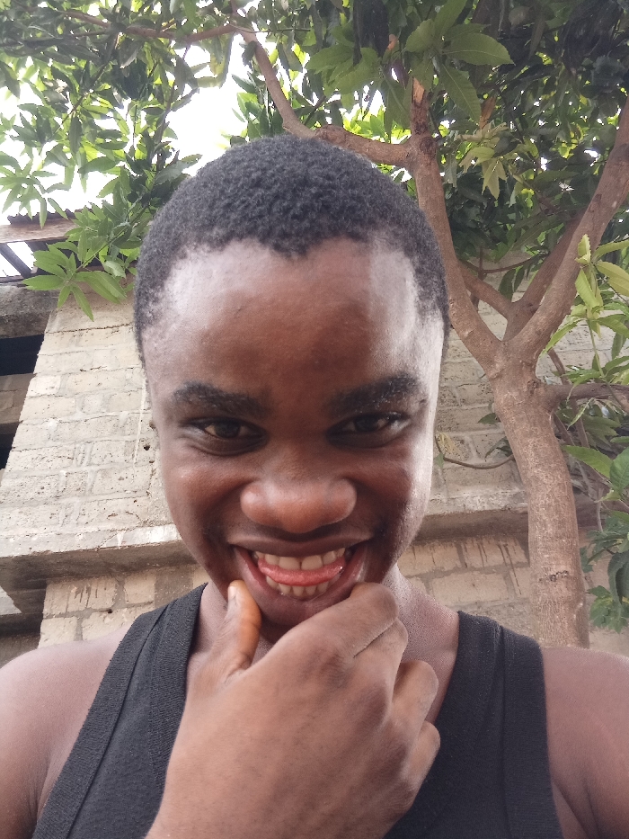 The image depicts a single individual in Owerri, Nigeria. The individual, with dark skin and short, dark hair, displays a broad smile with exposed teeth and a visible tongue, gazing directly at the viewer. Their right hand is positioned with fingers against the chin and mouth area. They are dressed in a black sleeveless garment. The scene is outdoors under diffused light. The background features a light-colored brick wall structure with a section of corrugated metal roofing to the left of the individual, and a tree with green foliage and visible branches to the right.