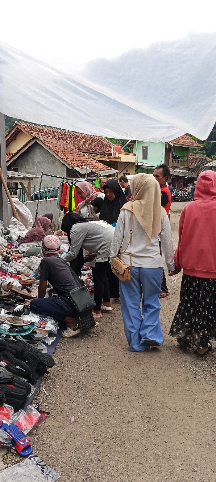 A group of people are gathered in front of a stall in the town of Kutawaringin, Indonesia, located at coordinates -6.974 latitude, 107.519 longitude. The stall is covered by a white tarp. The people are looking at and possibly purchasing shoes and sandals that are displayed on the ground. Several bags and other items are scattered around the stall. The people are dressed in a variety of clothing, including brightly colored shirts, long pants, and head scarves. Some are wearing backpacks. There are several houses and other buildings in the background, including a large green building with a balcony.