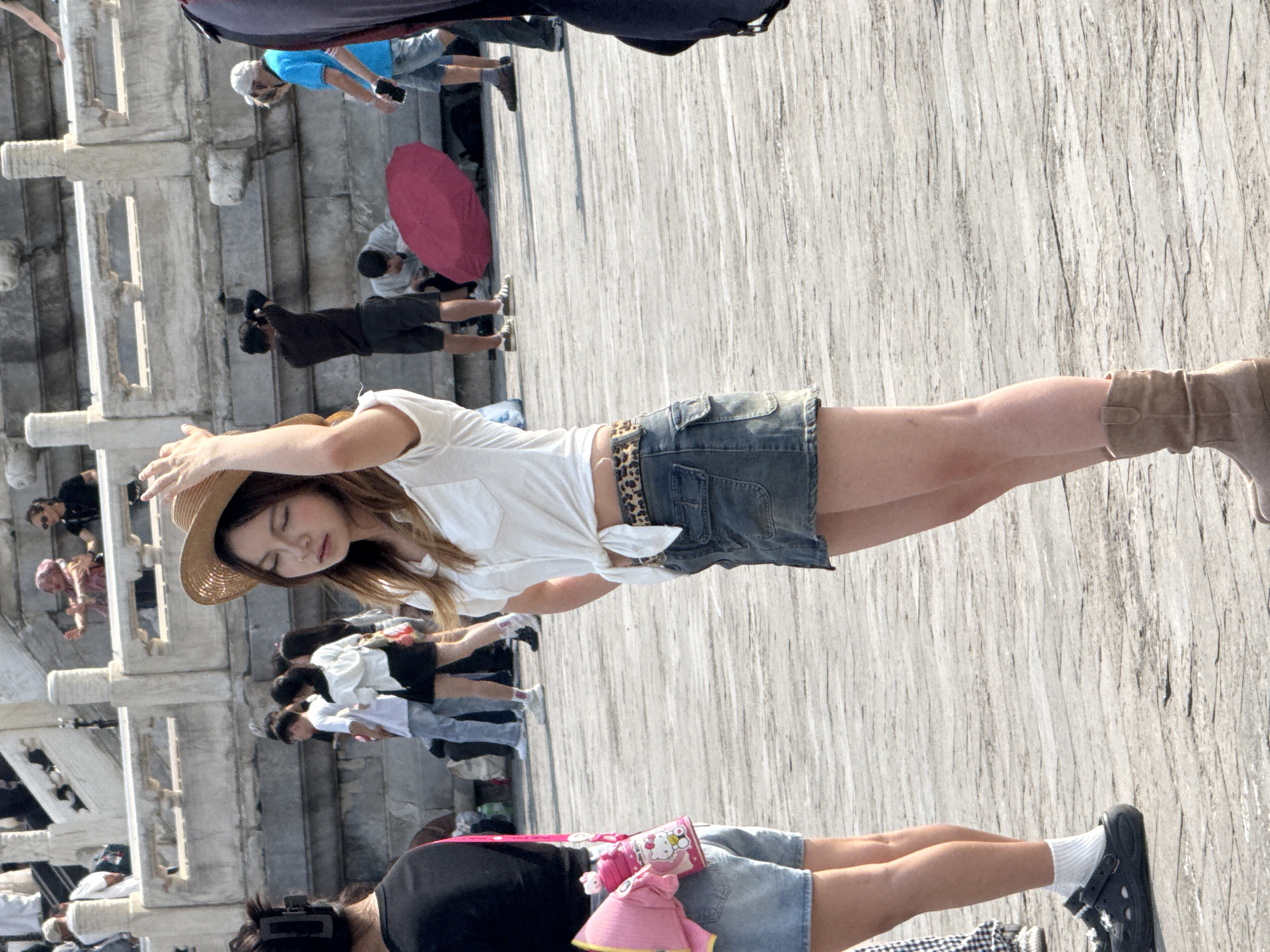 In Dongcheng District, China, a young woman wearing a straw hat, white short-sleeved shirt tied at the waist, and a denim mini-skirt stands on a paved walkway. She is facing sideways with her head tilted back and her eyes closed. Her right hand is raised and touching her hat. She is wearing tan ankle boots. To her left, another person wearing a black top and denim shorts is visible from the waist down. A pink Hello Kitty water bottle is clipped to their belt. In the background, several other people are walking and standing around, with stone architecture and steps visible. A bright red umbrella lies on the ground near some of the background figures. The scene appears to be a public outdoor space with historical architecture.