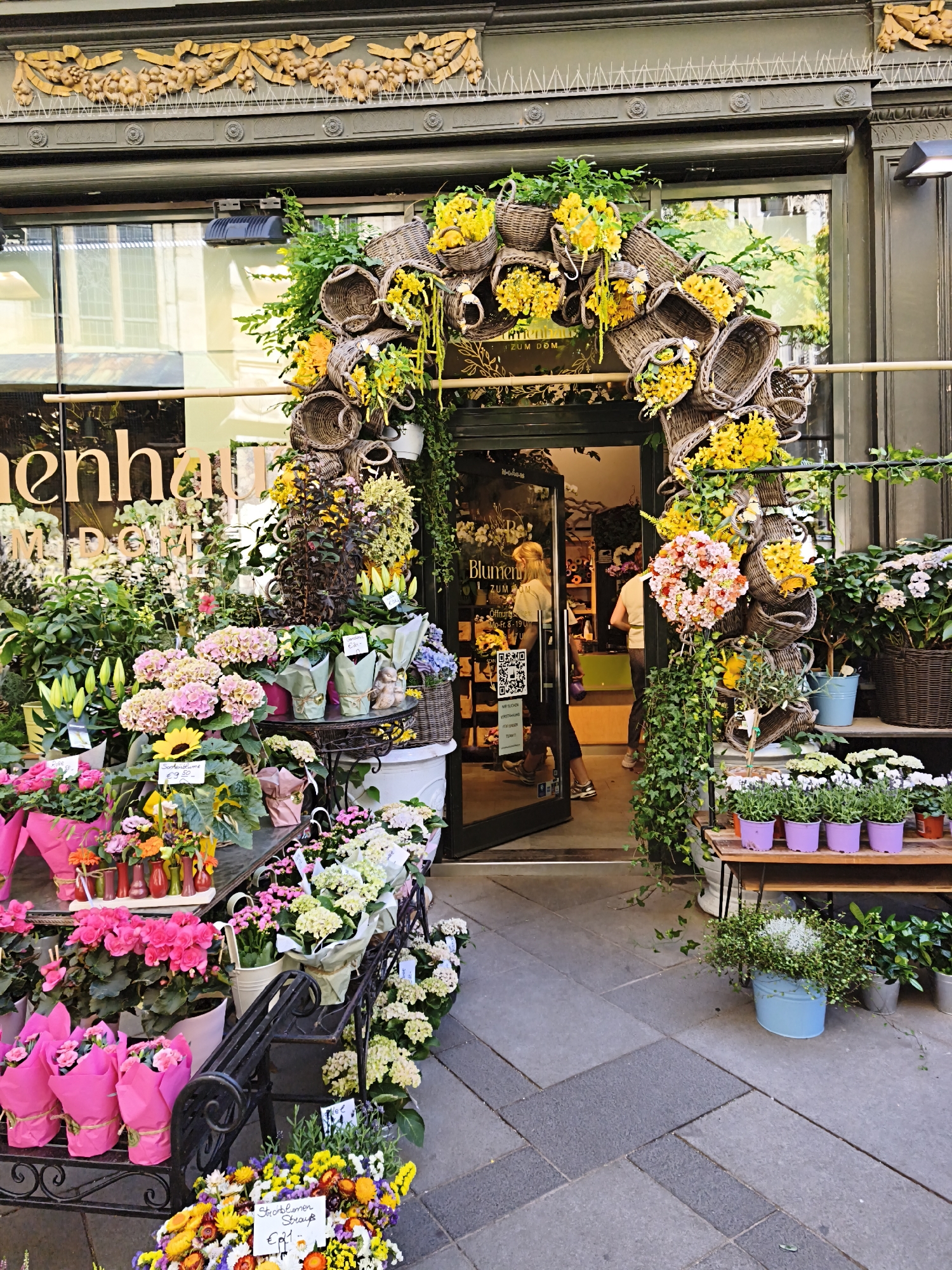 The image depicts the exterior of a flower shop named "Blumen ZUM DOM" located in Vienna, Austria. The shop's entrance is adorned with an archway constructed from woven baskets filled with yellow flowers and greenery. Various potted plants and bouquets of flowers are displayed on the sidewalk in front of the shop. Two women are visible inside the shop, one of whom appears to be a customer. The shop front features large windows with the store's name visible. The sidewalk is paved with large, grey stone slabs.