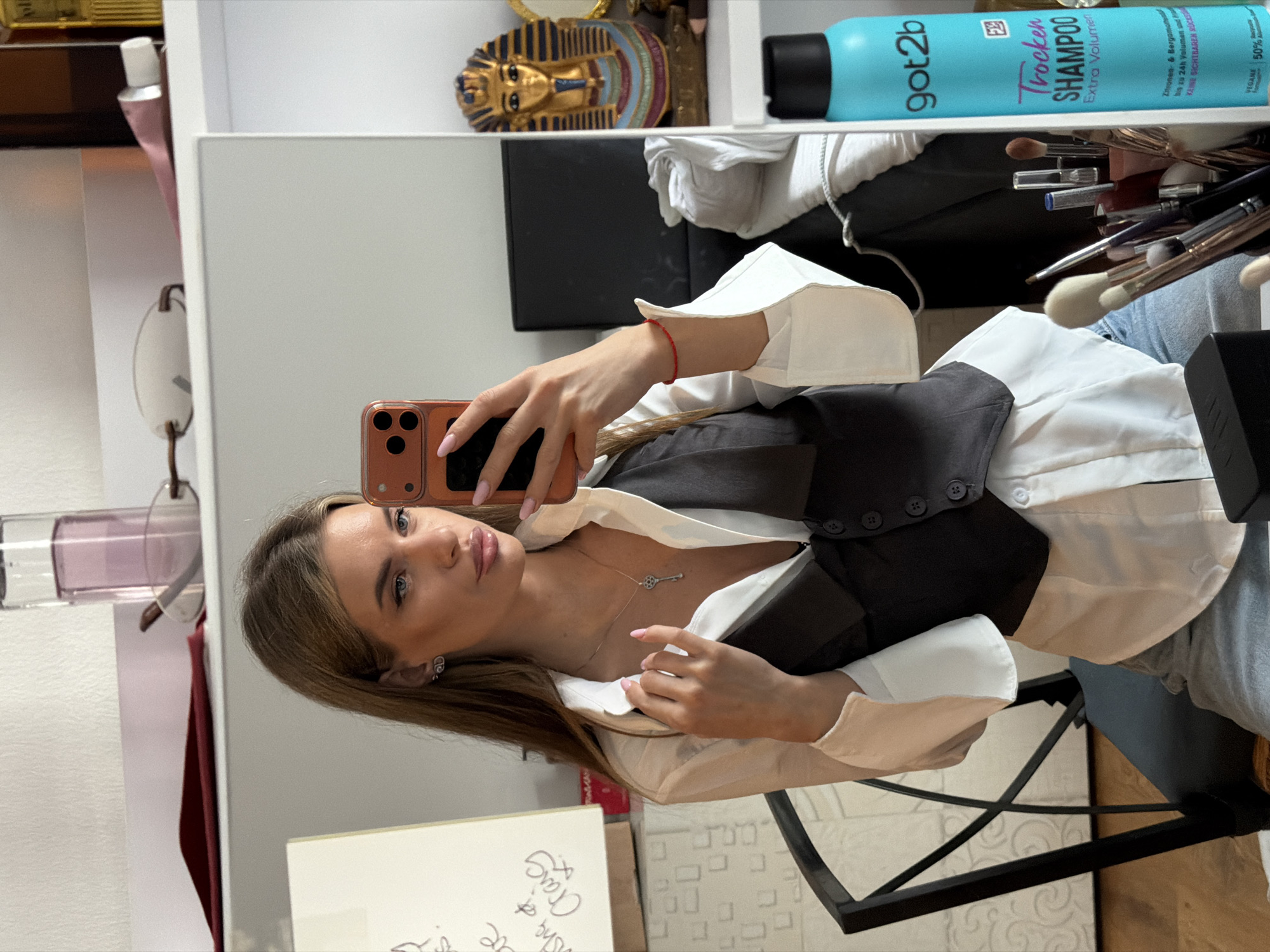 A woman is taking a selfie in a mirror. She is wearing a white button-down shirt with a gray vest. The mirror reflects a shelf with several items, including a bottle of "got2b Trocken SHAMPOO Extra Volumen," a makeup brush holder with brushes, and a small pharaoh figurine. The background appears to be a room interior. There's no evidence to indicate the location is Berlin, Germany.