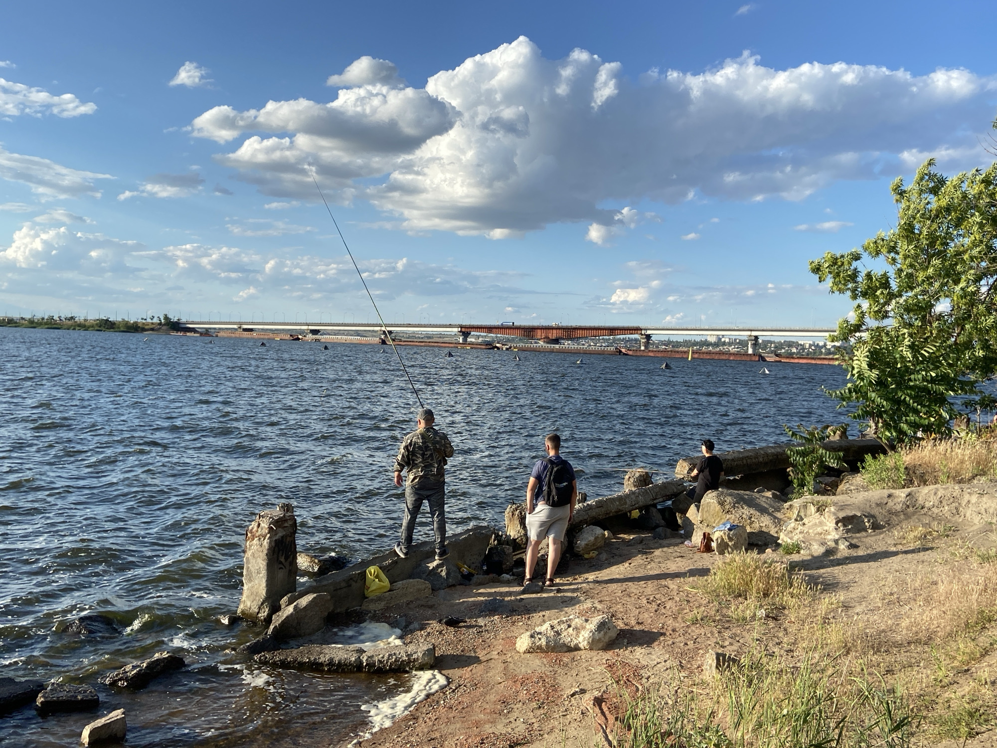 Two men are fishing on a rocky shore in Unknown, Unknown. One man, wearing a camouflage shirt and gray pants, stands on a rock to the left, holding a fishing rod. The other man, in a blue shirt and light-colored shorts, stands to the right, facing away from the camera. A third person, wearing a black shirt and black pants, sits on a concrete structure in the background. The water is choppy with whitecaps. In the distance, a large bridge spans the water, and several barges are visible. The shore is covered in dirt, rocks, and dry grass, with scattered debris, including yellow plastic bags. A tree with green leaves is located on the right side of the image. The sky is blue with large, fluffy white clouds.