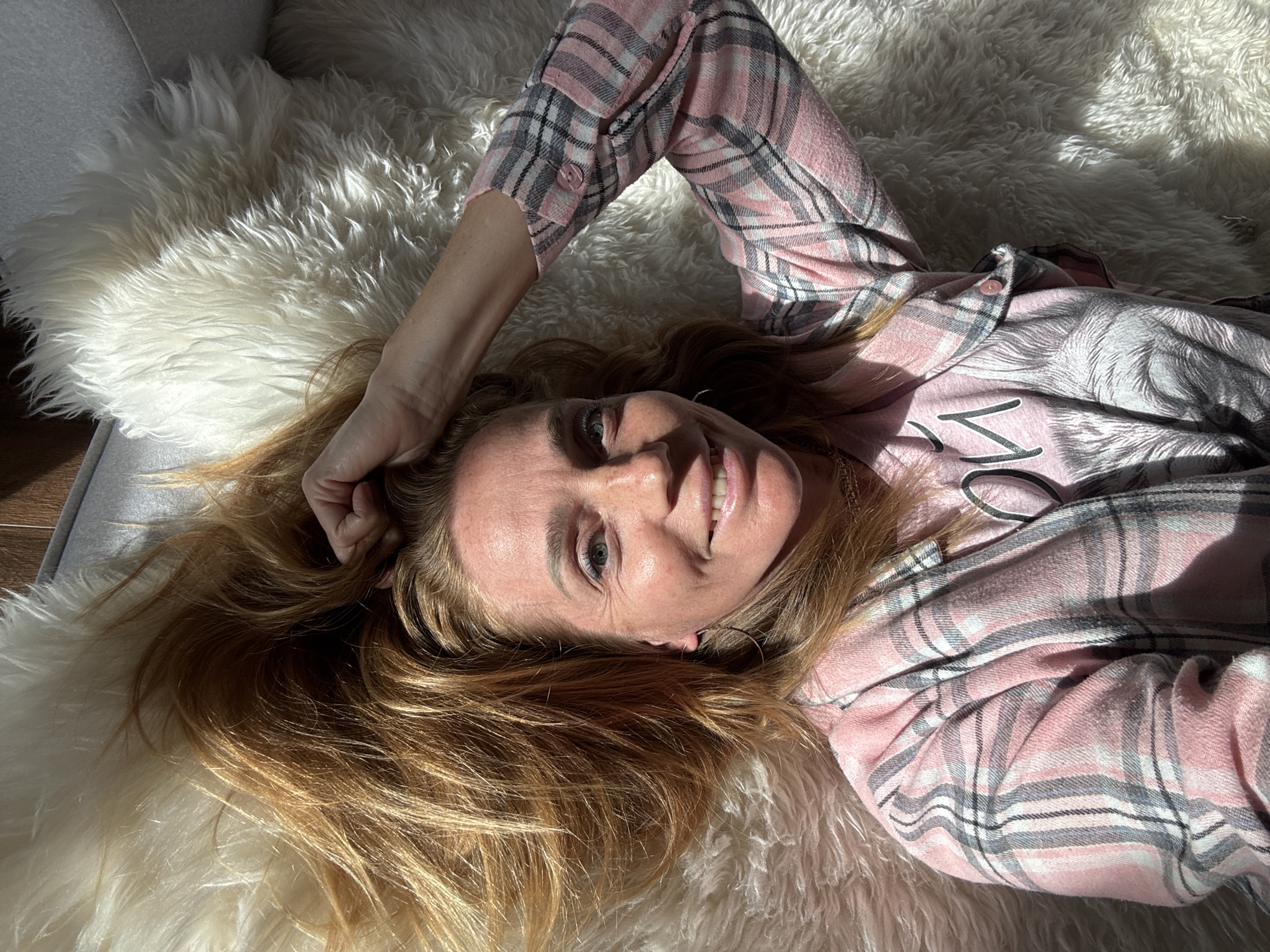 A woman with blonde hair lies on a white fluffy rug. She is wearing a pink plaid shirt. She has her right hand raised, resting on her head. The woman is smiling at the camera. Sunlight is casting shadows on her face and the rug. The background appears to be a wooden floor and a couch. There is no identifiable information indicating the location is Steklyannyi, Russia.
