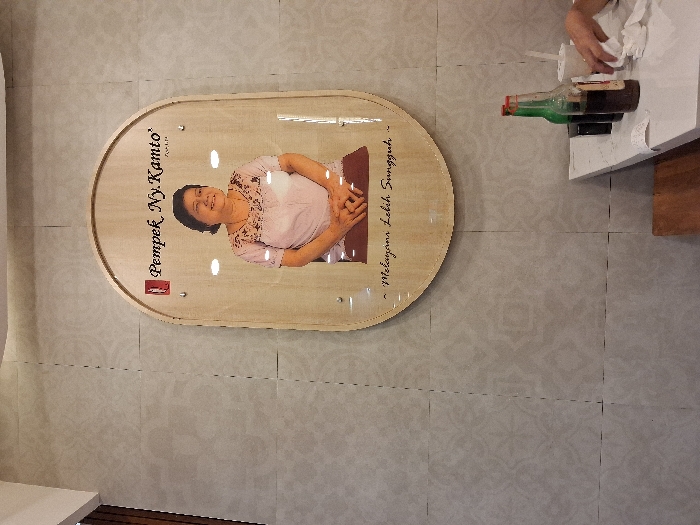 A light wood sign hangs on a wall with light-colored tiles. The sign is oval-shaped with a woman’s portrait, text reading “Pempek Ny. Kamto” and a line in Indonesian. At the top right, a person with a white cloth wipes a table. A bottle with a green neck and brown body sits on the table. A wooden object is under the edge of the table.