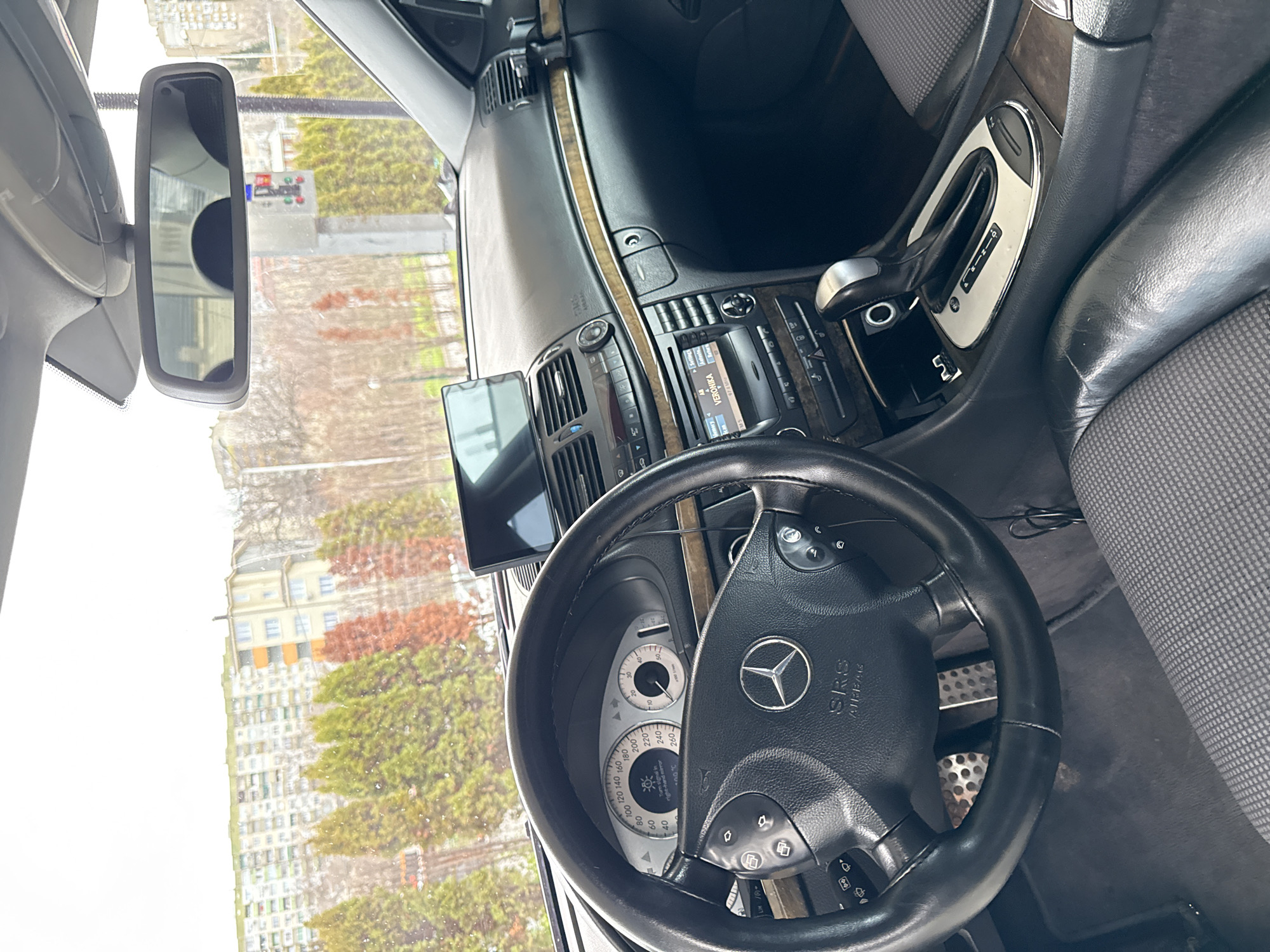 The image shows the interior of a Mercedes-Benz car, focusing on the driver's seat area. The steering wheel, dashboard, center console, and rearview mirror are visible. Outside the car, through the windshield and rearview mirror, one can see a city landscape with buildings and trees. The navigation screen shows "VERONIKA". The speedometer reads a temperature of 3 degrees Celsius. The gear stick is in "P" for park. Based on the architecture and foliage, the location appears to be Stara Zagora, Bulgaria. The rear view mirror and traffic lights suggest the vehicle is stationary.