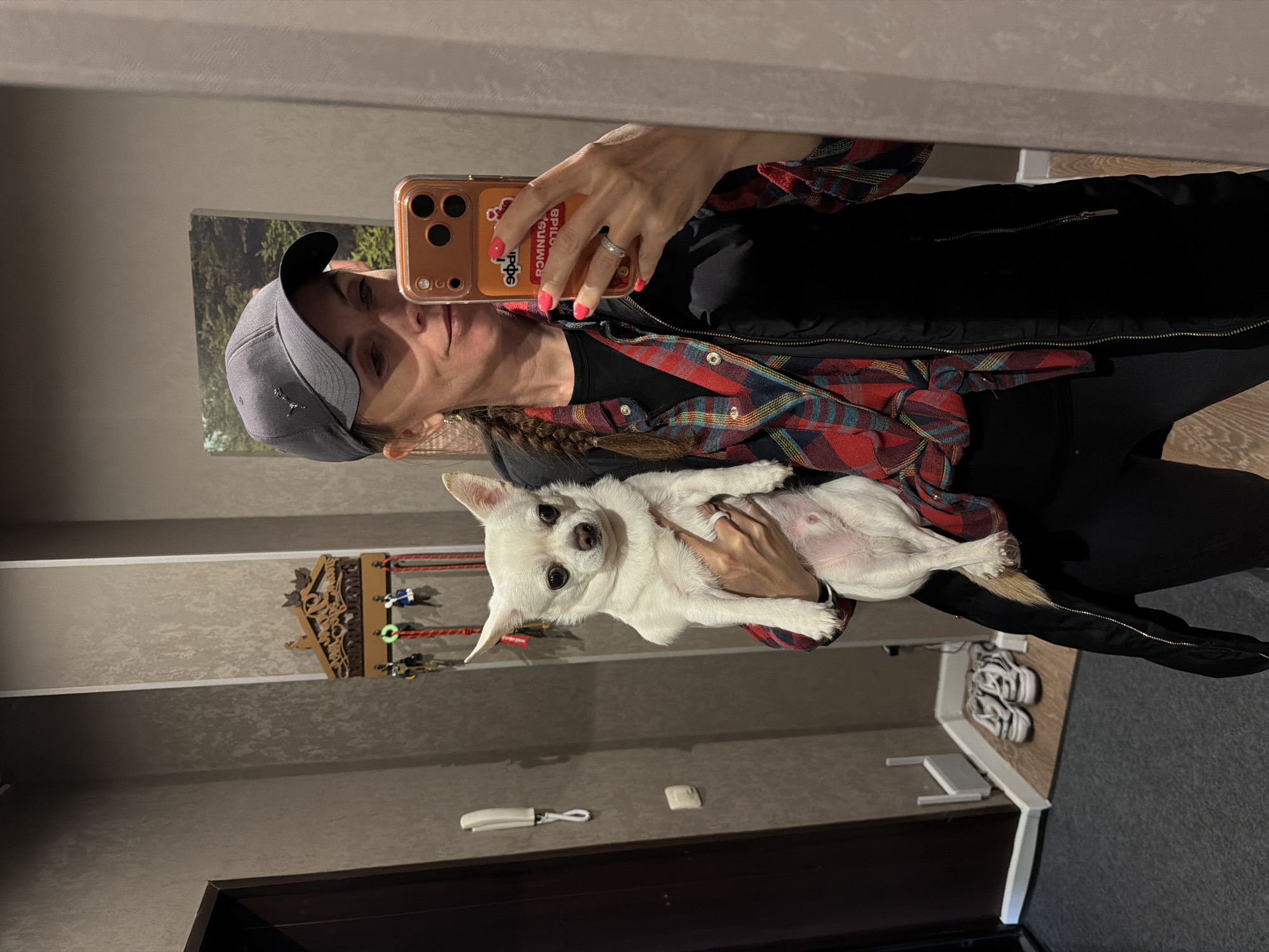 A woman in a gray baseball cap, black jacket over a red and black plaid shirt, and black pants is holding a small, white dog and taking a selfie in front of a mirror. The mirror reflects the room, which includes a wall decorated with a wooden wall hanging with hooks and leashes, a white electrical outlet, a white case, and a brown door frame. In the background is a gray carpet, a white table with two pairs of zebra striped sneakers underneath. The scene does not suggest the city of Birobidzhan, Russia; there are no indicators of location.