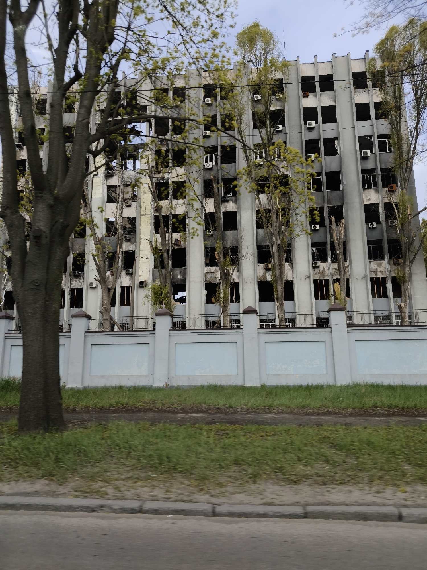 A building in Dnipro, Ukraine, stands heavily damaged. The gray, multistory structure has rows of empty, blackened window frames. The exterior is stained with soot. Air conditioning units are visible on some of the walls. A fence with gray posts and light blue panels runs along the front of the building. Trees with sparse green leaves stand between the fence and the building. A strip of green grass is in front of the fence, and a gray curb and paved road are in the foreground.