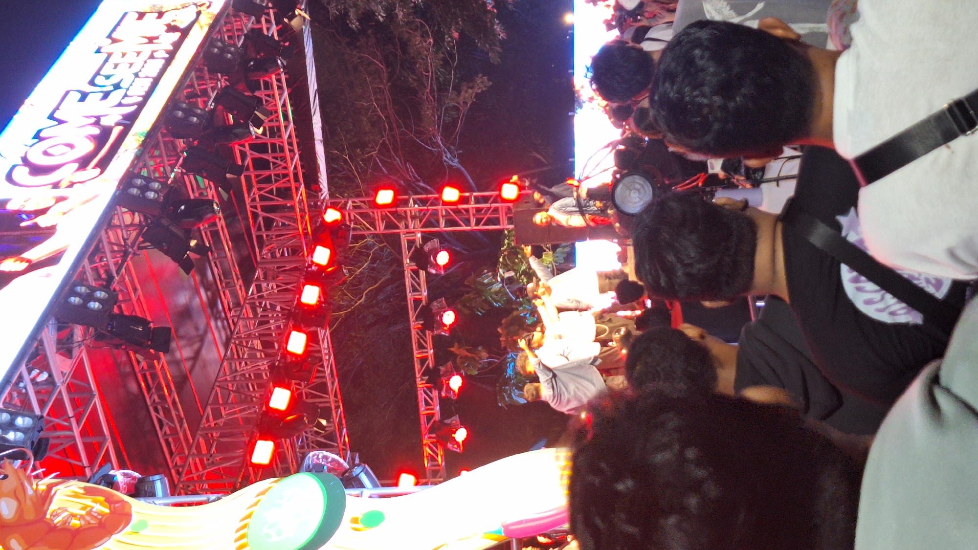 An outdoor concert stage is illuminated with red lights and stage lighting. A large screen displays text that reads "COME SEE ME". A crowd of people are visible in the foreground and midground, facing the stage. Several performers are on stage, some with their hands raised. The overall scene suggests a lively event taking place at night.