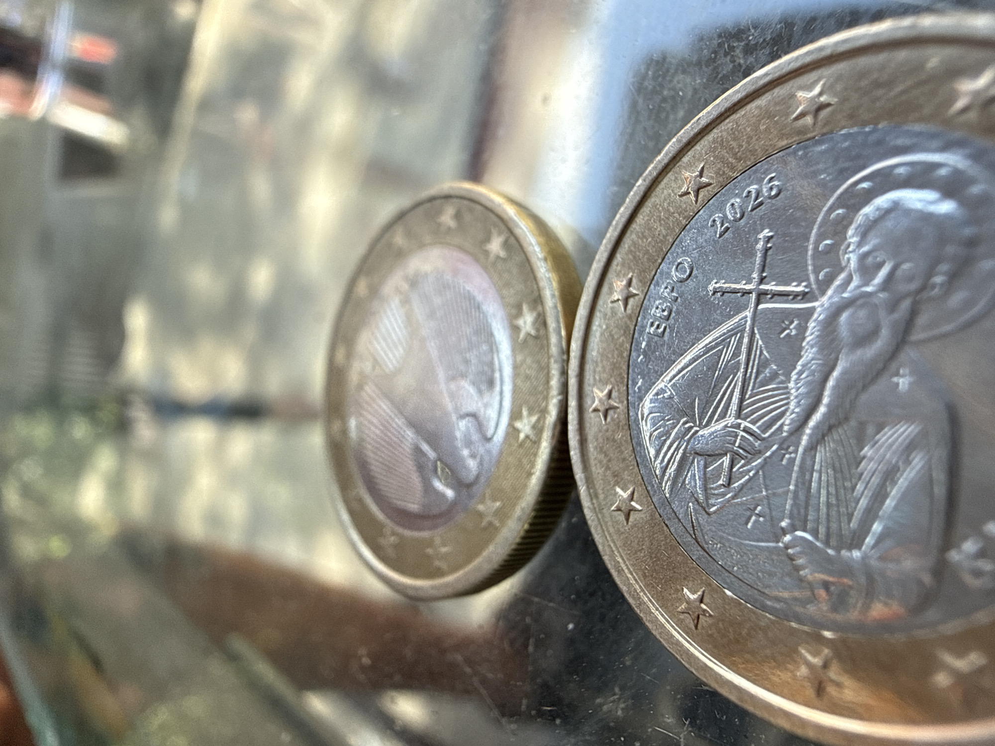The image displays two euro coins on a reflective surface, likely a glass tabletop, outdoors. The coin in the foreground is partially visible, showing a silver center with a golden outer ring. The design on this coin includes stars along the rim and text that appears to read "EURO 2025". The coin in the background is also partially visible, with a similar golden outer ring and a different design in its center, which is silver with some darker shading. The background is blurred but suggests an outdoor environment with greenery and possibly a paved area, with hints of sunlight filtering through. There are no people visible in the image, and the scene appears to be a close-up, still life shot of the coins. There is no information in the image to suggest a specific location within the city of Varna, Bulgaria.