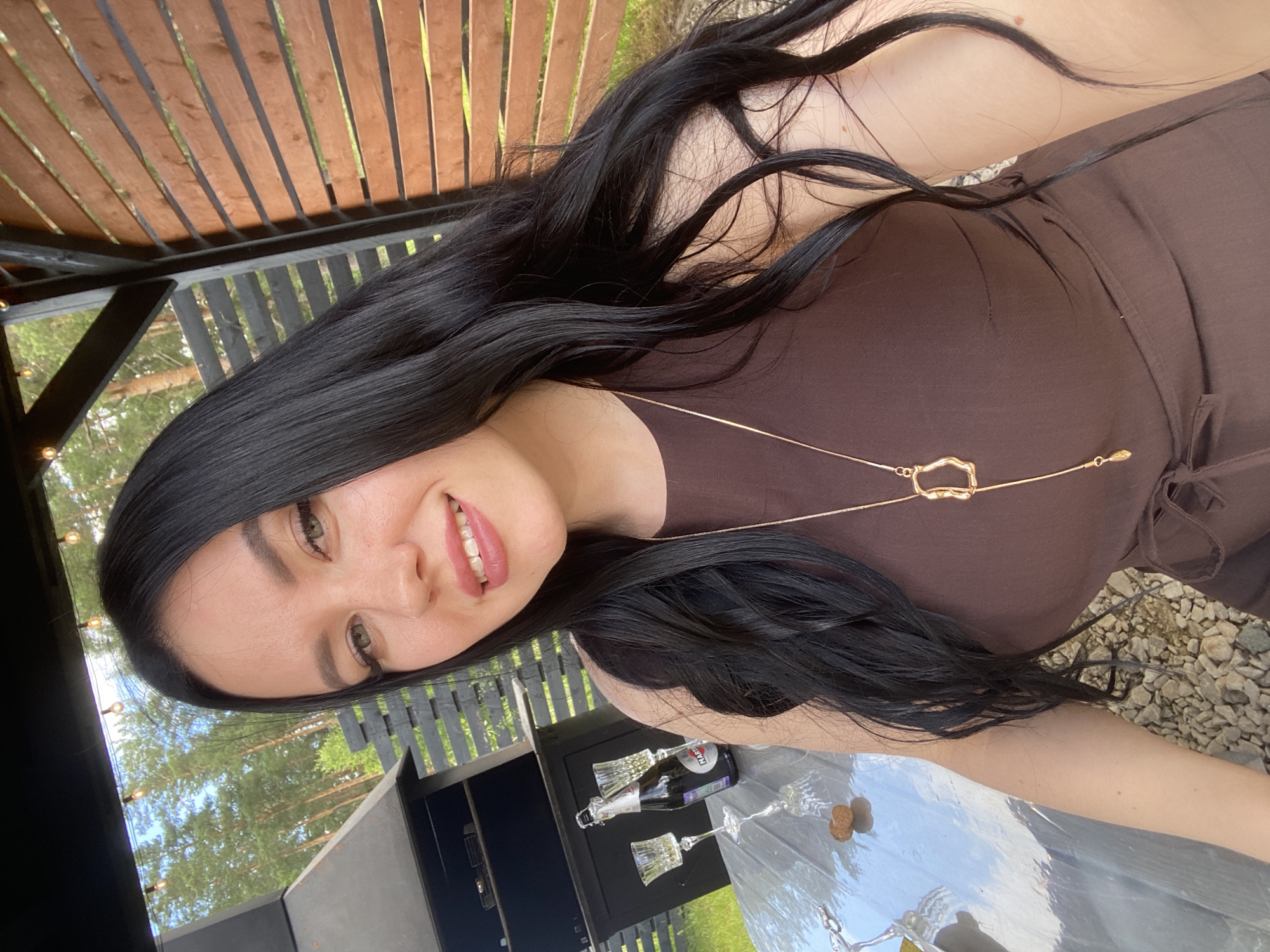 The image shows a woman with long, dark hair and light skin, wearing a brown sleeveless dress and a gold necklace with a pendant. She is outdoors, possibly in a backyard or patio area, with a wooden fence and trees in the background. There is a table with a glass bottle of champagne and two champagne flutes. A grill or outdoor kitchen area is visible to the left. The ground is covered with gravel and some scattered stones. The woman appears to be smiling and looking at the camera. The location is stated to be in the city of Chaikovsky, Russia.