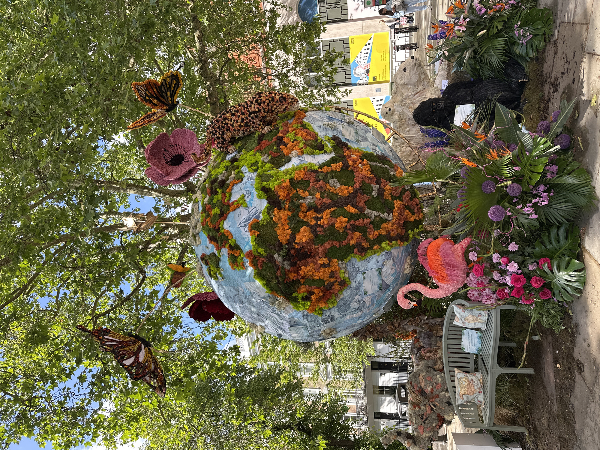 The image captures an outdoor scene in London, United Kingdom. A large globe sculpture, representing the Earth, dominates the foreground. The continents on the globe are rendered in green, orange, and brown moss-like material, with blue representing the oceans. Artificial flowers and a leopard figure are placed on top of the globe. Artificial butterflies and a flamingo are attached to the globe and the surrounding foliage.
To the right of the globe, a bench is situated amongst lush plants and flowers. Other floral sculptures of animals are nearby, positioned amongst additional vibrant flowers and tropical plants.
In the background, buildings are visible, along with a few people standing and walking by. The scene appears to be in a public park or garden setting within the city.