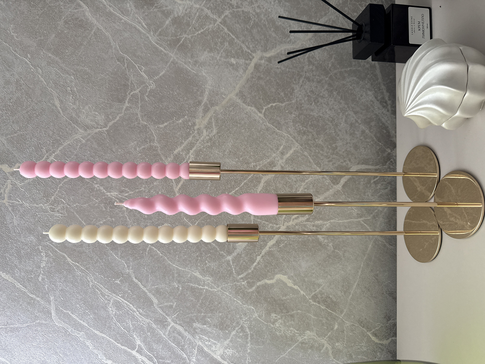 In Mahilyow, Belarus, on a white surface in front of a gray, marbled wallpaper, sit three tall, slender, gold-toned candle holders. Each candle holder supports a candle: the leftmost holds a cream-colored candle with a bubble-like texture, the middle holds a pink, twisted candle, and the rightmost holds a pink candle with a bubble-like texture. To the right of the candle holders, a black square diffuser bottle with black reeds stands. In the foreground, a white, shell-shaped decorative object is visible.