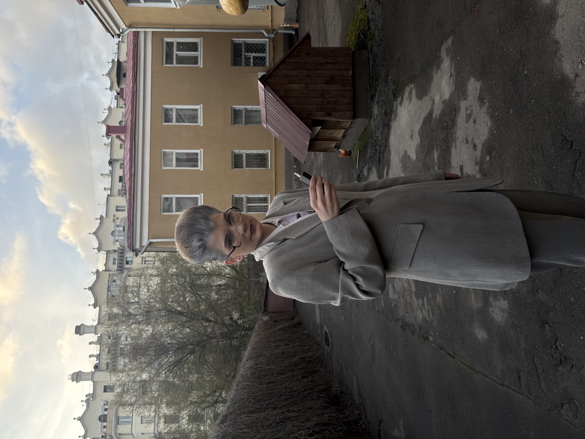 The image captures a person standing outdoors in an urban setting, likely a courtyard or alleyway in Moscow. The central subject is a person with short, grayish-blonde hair, wearing dark-framed glasses, a light grey or beige suit jacket, and a white shirt. They hold a small, dark cylindrical object, possibly a vape pen, in their right hand and look directly towards the camera with a neutral expression.
In the background, several buildings are visible. Directly behind the person is a prominent, light brown or tan multi-story building with numerous rectangular windows. To the left and further in the distance, another, lighter-colored building features more ornate architectural details along its roofline, including decorative elements and chimneys. A small, dark brown wooden shed with a reddish-brown roof stands behind the person, towards the right.
The foreground shows asphalt pavement that appears worn, with visible cracks and patches. On the left side of the frame, the bare branches of a tree indicate a colder season, such as late autumn, winter, or early spring. A dense, low bush or hedge also lines the left side of the foreground.
The lighting suggests late afternoon or early evening, with a soft, diffused light and a partly cloudy sky. The overall atmosphere is quiet and still. There is no visible text in the image.