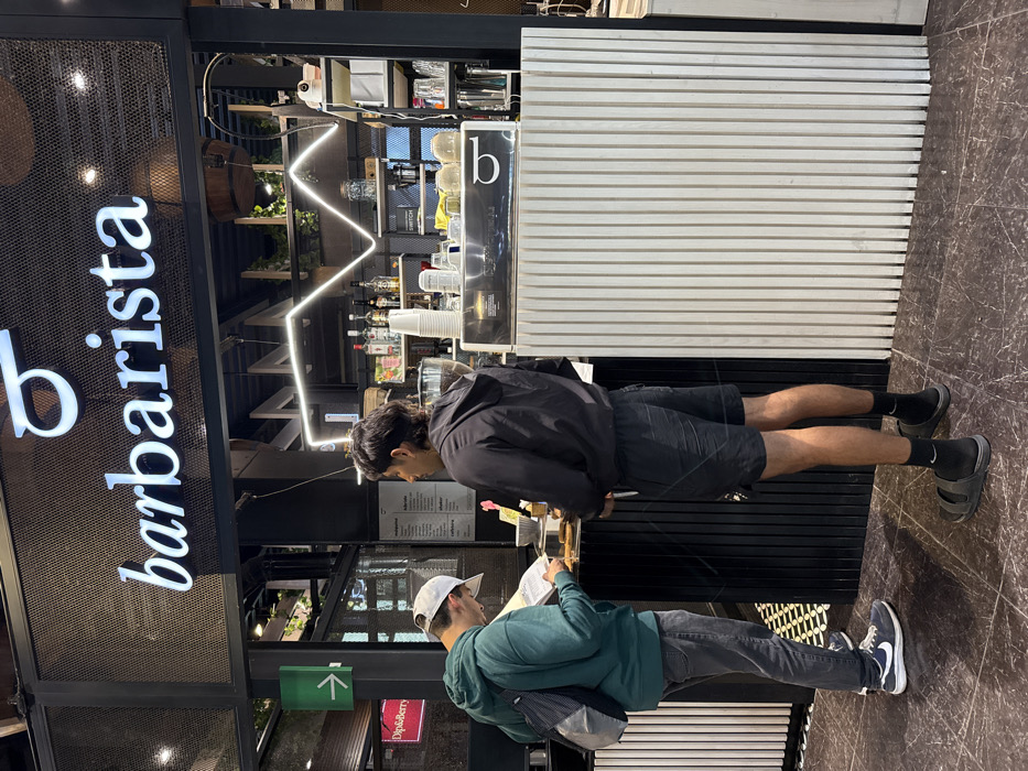 Two men stand at a counter inside a "barbarista" cafe. The first man wears a black jacket, shorts, and black socks with sandals. He looks down at something on the counter. The second man wears a white baseball cap, a green jacket, gray jeans, and blue sneakers, and reads a menu. Behind them, shelves display various items and a zigzag neon light fixture decorates the wall. The cafe's name is displayed prominently in neon letters.