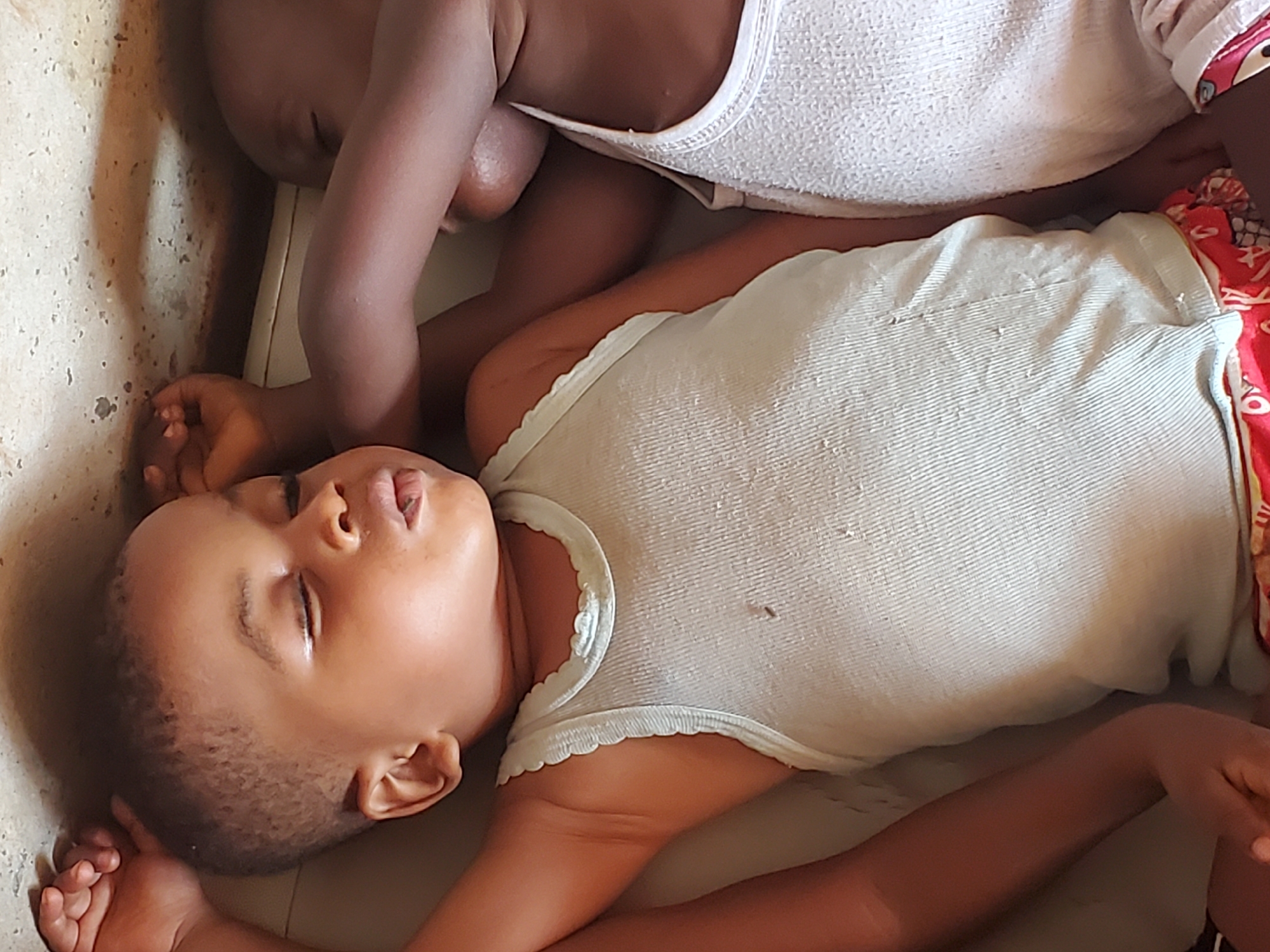 In Ukwu-Abwa, Nigeria, two young children sleep on a surface. One child wears a white ribbed tank top and printed shorts. The second child wears a white tank top. The surface appears to be adjacent to a wall with textured stucco.