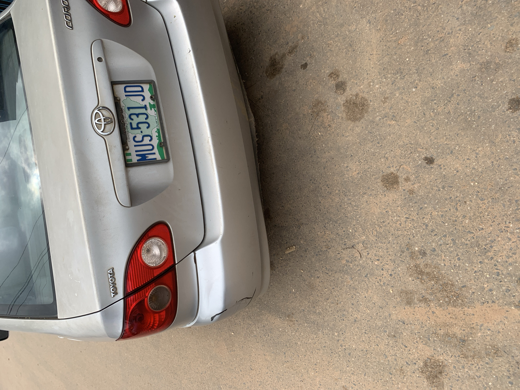 The image shows the rear of a silver Toyota Corolla car parked on a dirt road. The car has a Nigerian license plate that reads "MUS-531 JD". The car has some damage to the rear bumper. The environment appears to be in a dusty, urban area. Based on the license plate and the style of the car, the image is likely taken in Nigeria. Given that the license plate is from Nigeria and the car is a Toyota Corolla, a common vehicle in Nigeria, and considering the context provided, the scene is likely located in Shagamu, Nigeria.