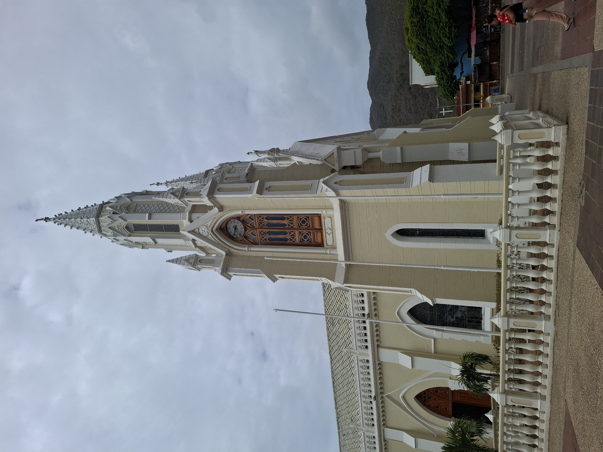 This media file captures a detailed view of a prominent church building in El Valle del Espíritu Santo, Venezuela, on an overcast day.
The central subject is an ornate church, likely the Basílica Menor Nuestra Señora del Valle, characterized by its cream-colored walls and intricate white architectural details. It features a tall, multi-tiered spire at the front, adorned with finials and decorative elements. A clock face with Roman numerals is visible on the main tower. The church exhibits a Neo-Gothic architectural style, evident in its pointed arches, decorative windows, and elaborate balustrades.
In the foreground, a white balustrade runs parallel to the church, separating it from a paved area or sidewalk. Small green plants are visible near the church's base and behind the balustrade. A flagpole stands prominently near the front. On the far right, two people are partially visible, one wearing light trousers and a dark top, holding a red object, possibly a bag, while walking on the paved surface.
The background features a large, dark mountain on the right, contributing to a scenic setting. Lush green foliage is also visible, indicating trees or other vegetation. The sky is uniformly overcast with light grey clouds, suggesting a cloudy daytime. The image focuses on the architectural beauty and surrounding environment of this significant religious structure.