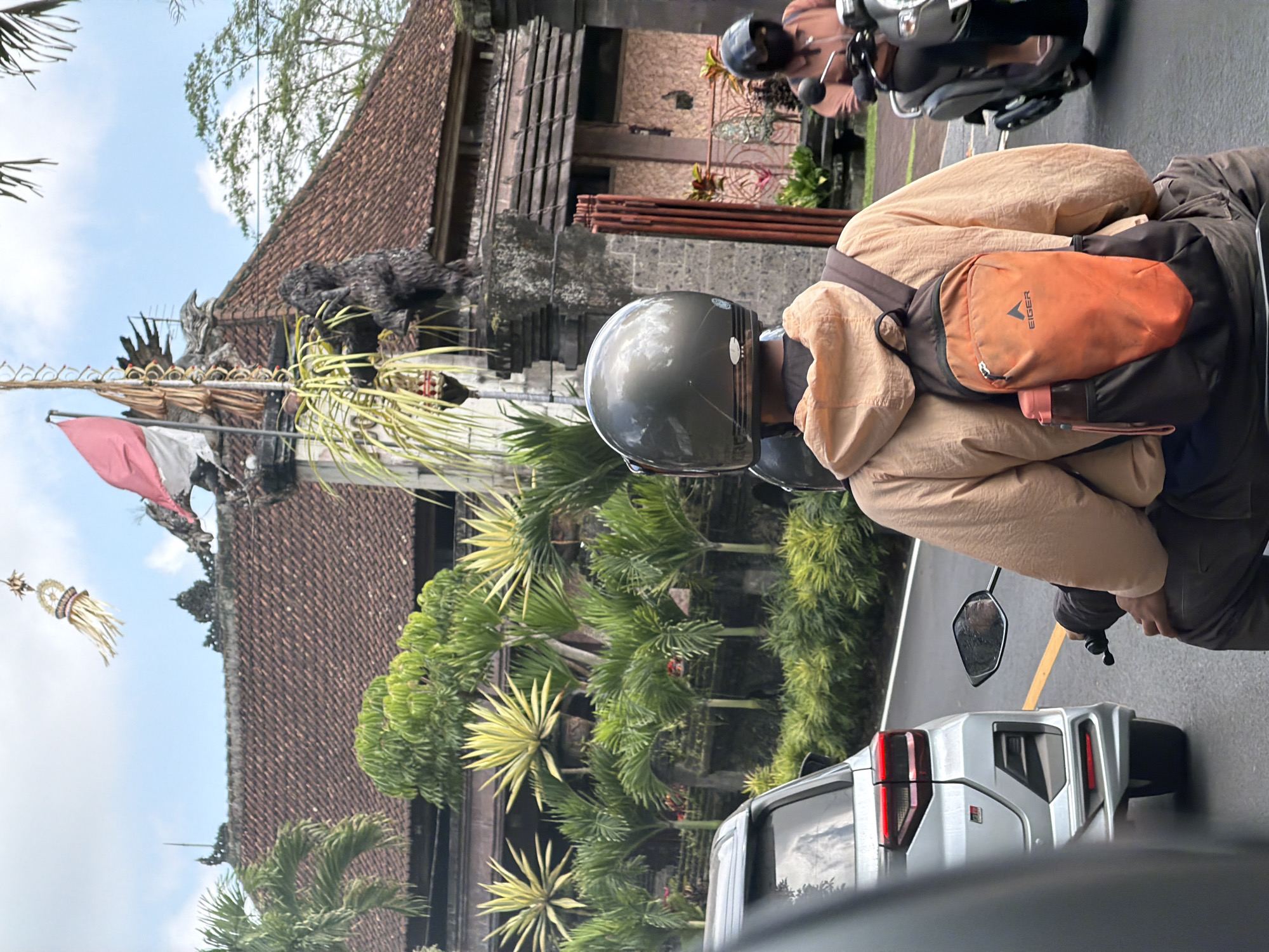 A medium shot captures a person riding a motorcycle in Tampaksiring, Indonesia. The rider is wearing a dark grey helmet and a light brown hooded jacket over dark pants. A dark backpack with an orange pouch featuring the Eiger logo and a water bottle is strapped to their back. To the left, the rear of a silver car is visible. To the right, another person on a scooter wears a black helmet and a light brown shirt. Behind the motorcyclists, a building with a red tile roof and ornate stonework is prominent, adorned with tropical plants and a flagpole flying a red and white flag. The scene is set against a backdrop of lush greenery and a cloudy sky.