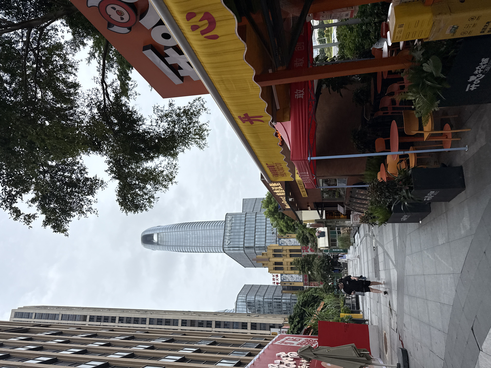 A street scene in Haizhu District, China features a pedestrian walkway lined with buildings and food stalls. On the left, a tall, modern building with many windows dominates the background. To its right, another contemporary building and a shorter, yellowish building with arched windows are visible. In the foreground, a street artist's stall with a red canopy and yellow awning displays food items. Orange and yellow tables and chairs are set up outside the stall. A few people are walking in the distance on the paved walkway. The sky is overcast.