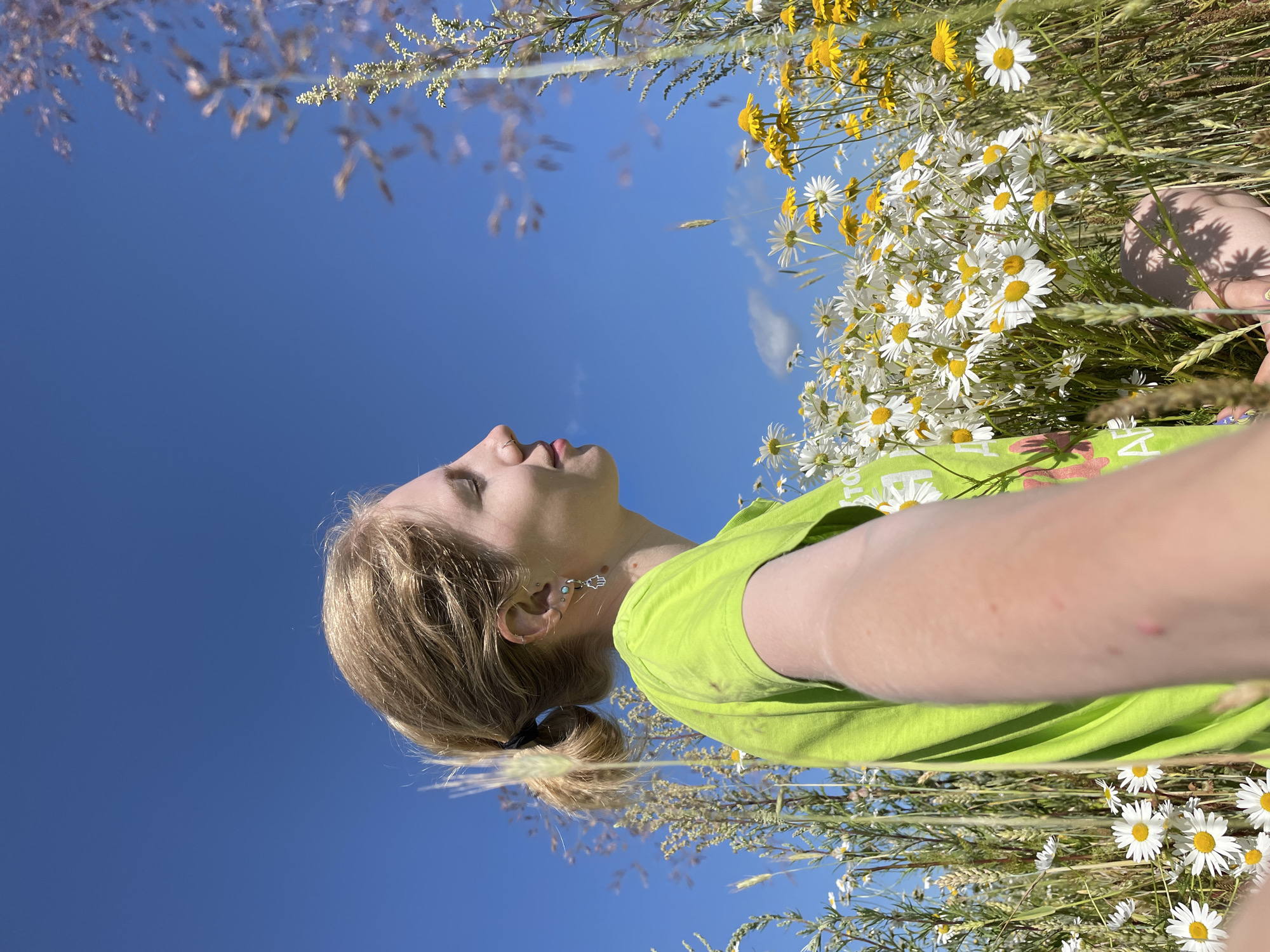 In the city of Сям-Можга, Russia, a young woman with blonde hair pulled into a ponytail, wearing a bright green t-shirt and earrings, is captured in a selfie. She is surrounded by a field of white and yellow wildflowers under a clear blue sky. The woman is shown from the shoulders up, facing to the right with her eyes closed and a slight smile. Her left arm is extended towards the camera, holding a bouquet of the wildflowers she is amidst. The background is dominated by the vibrant blue sky and the lush green and yellow hues of the meadow, with some taller grasses framing the scene.