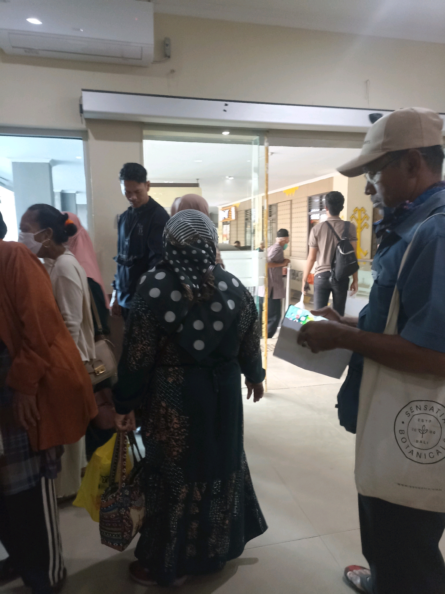 In Bandar Lampung, Indonesia, a group of people are gathered in what appears to be a hospital or clinic waiting area. An air conditioning unit is mounted on the wall above a large sliding glass door. Several individuals are seen from behind, including a person wearing a black hijab with white polka dots and a patterned dress. Another person, wearing an orange top and black pants with white stripes, is partially visible. A man in a light blue shirt and a beige baseball cap is in the foreground, holding a document and a white tote bag with a circular logo that reads "SENSATIA BOTANICAL ESTD 2020 BALI." Other individuals in the background include a man in a black jacket and others wearing masks and casual attire. The environment suggests an indoor public space with tiled flooring.