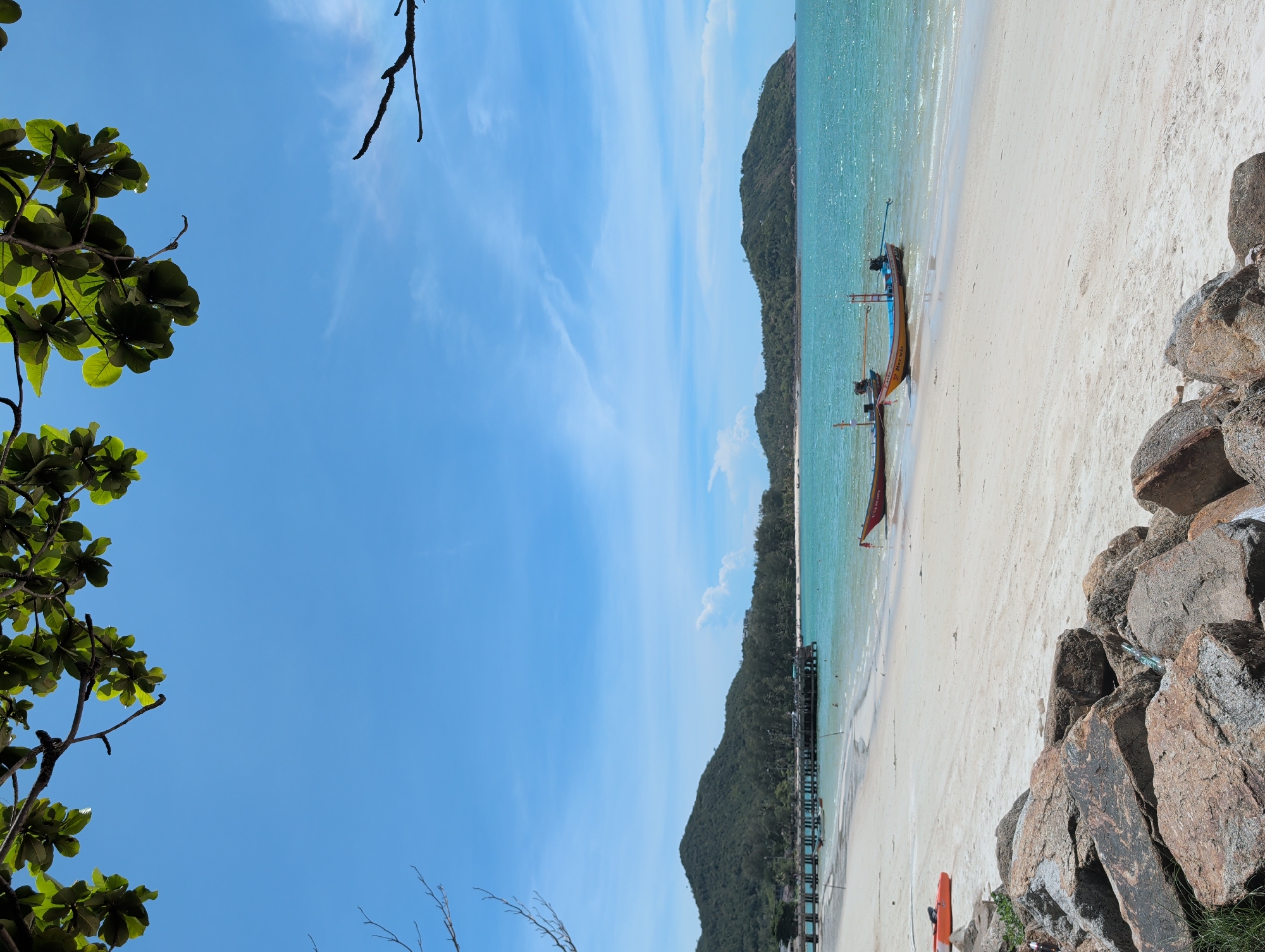 The image captures a scenic view of a beach in ตำบลเกาะพะงัน, Thailand. The sky is a clear blue with light clouds. The turquoise water of the sea meets the white sandy beach. Two traditional Thai longtail boats are docked on the shore. Lush green vegetation covers the hills in the background. In the foreground, rocks are visible on the right side, and tree branches with green leaves are present on the left. A long pier extends from the beach into the water.