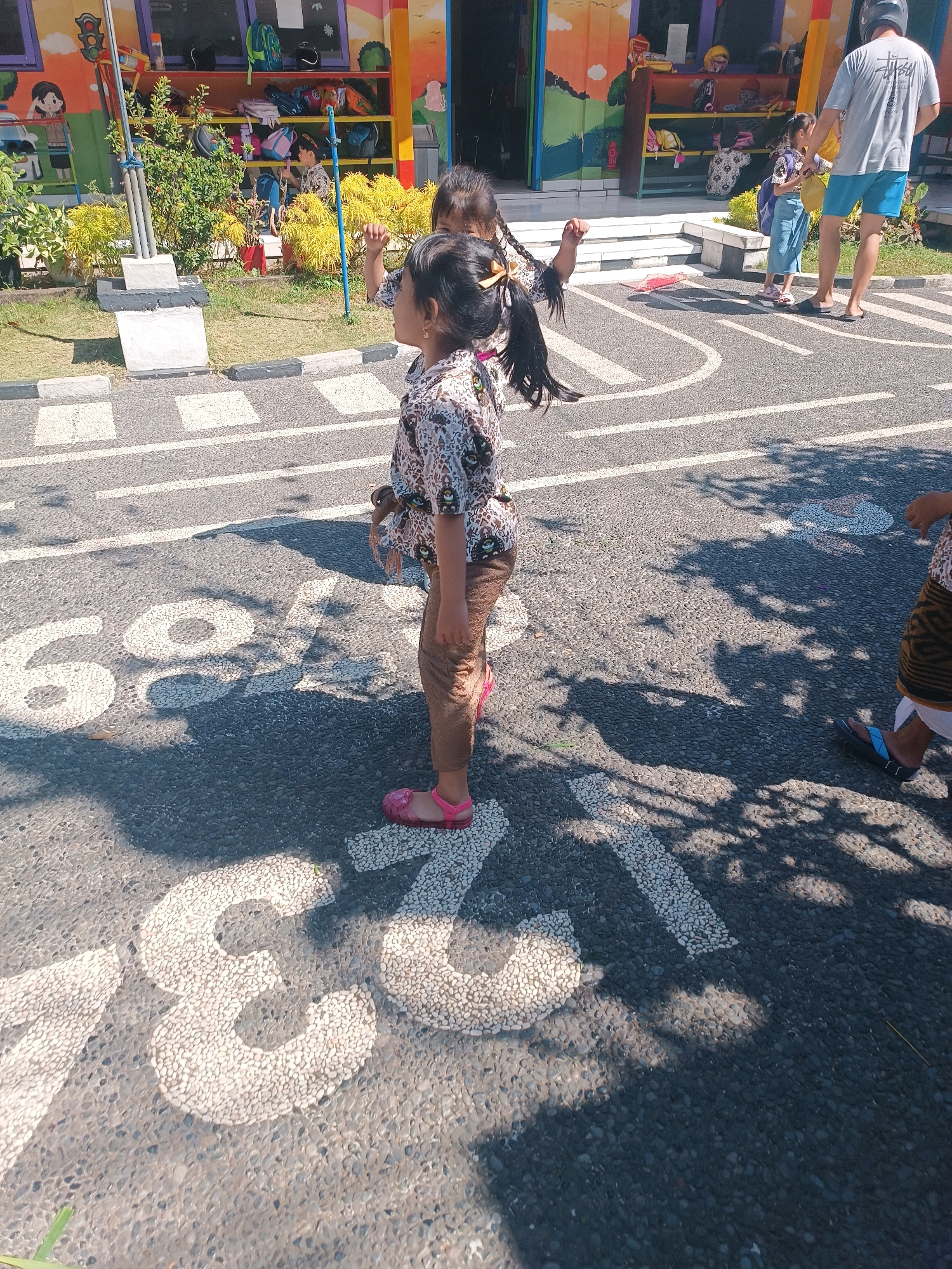 In Tabanan, Indonesia, a girl with dark hair in braids, wearing a patterned shirt and brown pants, stands on an asphalt surface marked with white painted numbers and lines. She is facing right, with her left arm bent and hand raised. Behind her, another child and a man wearing a grey t-shirt and blue shorts are visible, with a building in the background. The building features colorful murals and shelves stocked with items. The environment appears to be outdoors, possibly a schoolyard or playground, with shadows from trees cast on the ground.