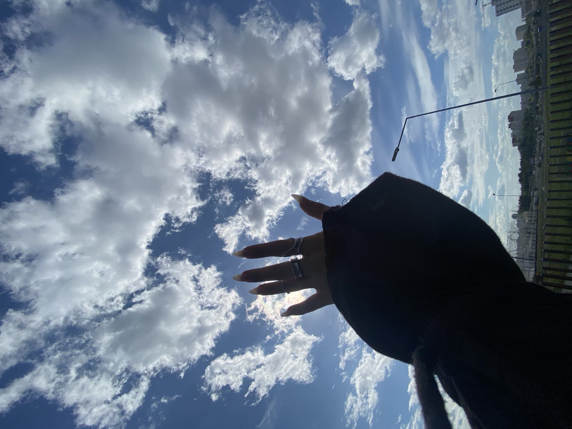 A hand, adorned with rings, reaches up towards a cloudy blue sky. The hand is silhouetted against the sun. Below the sky, a cityscape is visible, with buildings and streetlights indicating an urban environment. In the foreground, a dark sleeve obscures the rest of the person. The image appears to be taken in Kudrovo, Russia, given the specific architectural style of the buildings and the presence of streetlights commonly found in the region.