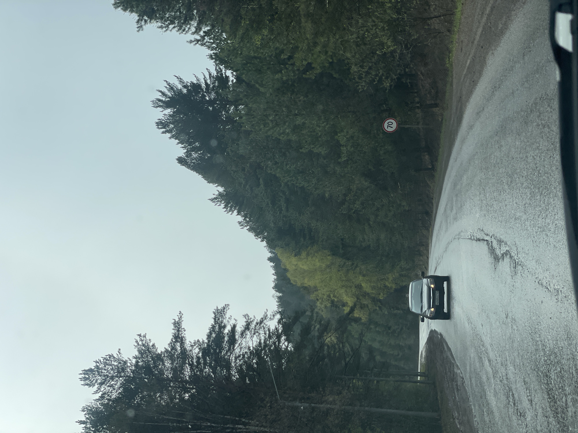 A vehicle travels on a wet road in Манский, Russia. The road curves to the right, bordered by dense trees. A circular road sign marked "70" is visible on the roadside. The sky is overcast.