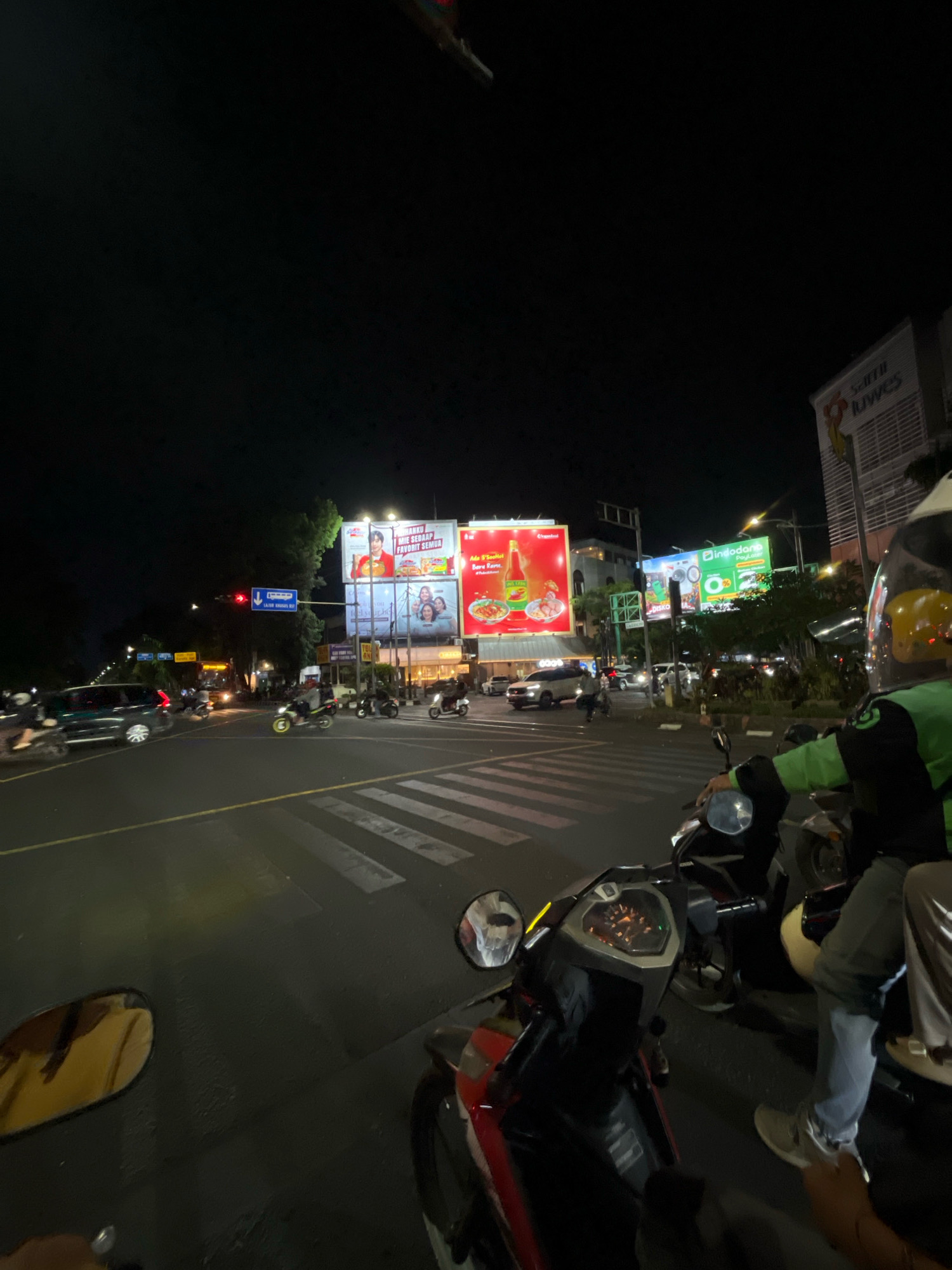 This is a street-level, night-time photograph taken from the perspective of someone on a motorcycle at an intersection in Surakarta, Indonesia. The scene is dominated by the darkness of night, punctuated by the bright lights of advertising billboards, streetlights, and vehicle headlights.
In the foreground, the front of a motorcycle is visible, with its instrument panel and handlebars in focus. A rider, wearing a green jacket and helmet, is on a motorcycle to the right of the frame. Another person, also on a motorcycle, is partially visible in the lower right corner.
The intersection itself is marked with pedestrian crosswalks and traffic lines. Cars and motorcycles are navigating the intersection. One car, a dark SUV, is visible on the left side of the frame.
Dominating the background are several large, illuminated advertising billboards. One billboard on the left displays an advertisement for "Mie Sedaap" instant noodles, featuring images of a man and two women. Another billboard in the center advertises "Indodana PayLater" with a green and white design. To the far right, a building with a large sign displaying "SULTAN" and "JUWES" is visible.
The presence of the advertising billboards, the Gojek (ride-hailing service) jacket on one of the riders, and the general urban night scene are characteristic of a bustling commercial area in Surakarta. The overall impression is one of urban life continuing into the night, with traffic and commerce under the glow of artificial lights.