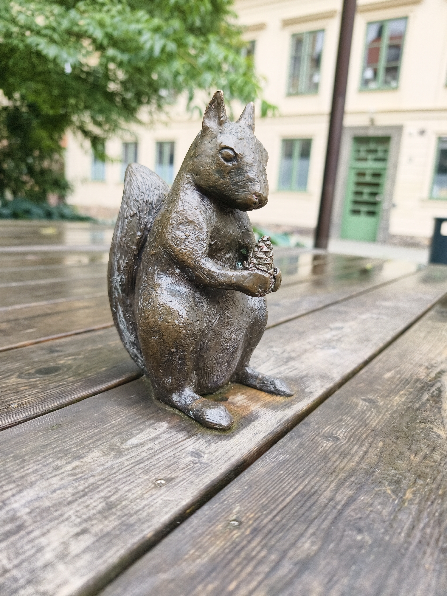 A bronze statue of a squirrel is positioned on a weathered wooden picnic table. The squirrel is depicted sitting upright, holding a pinecone in its paws. The table's surface is damp, suggesting recent rainfall. In the background, a light-colored building with green-trimmed windows and a green door is visible. Lush green trees flank the building. A trash can can be seen to the right of the building. The scene appears to be an outdoor seating area in a city. The statue is located in Gamla Linköping, a historical open-air museum in Linköping, Sweden, which preserves and showcases old wooden buildings from the region.