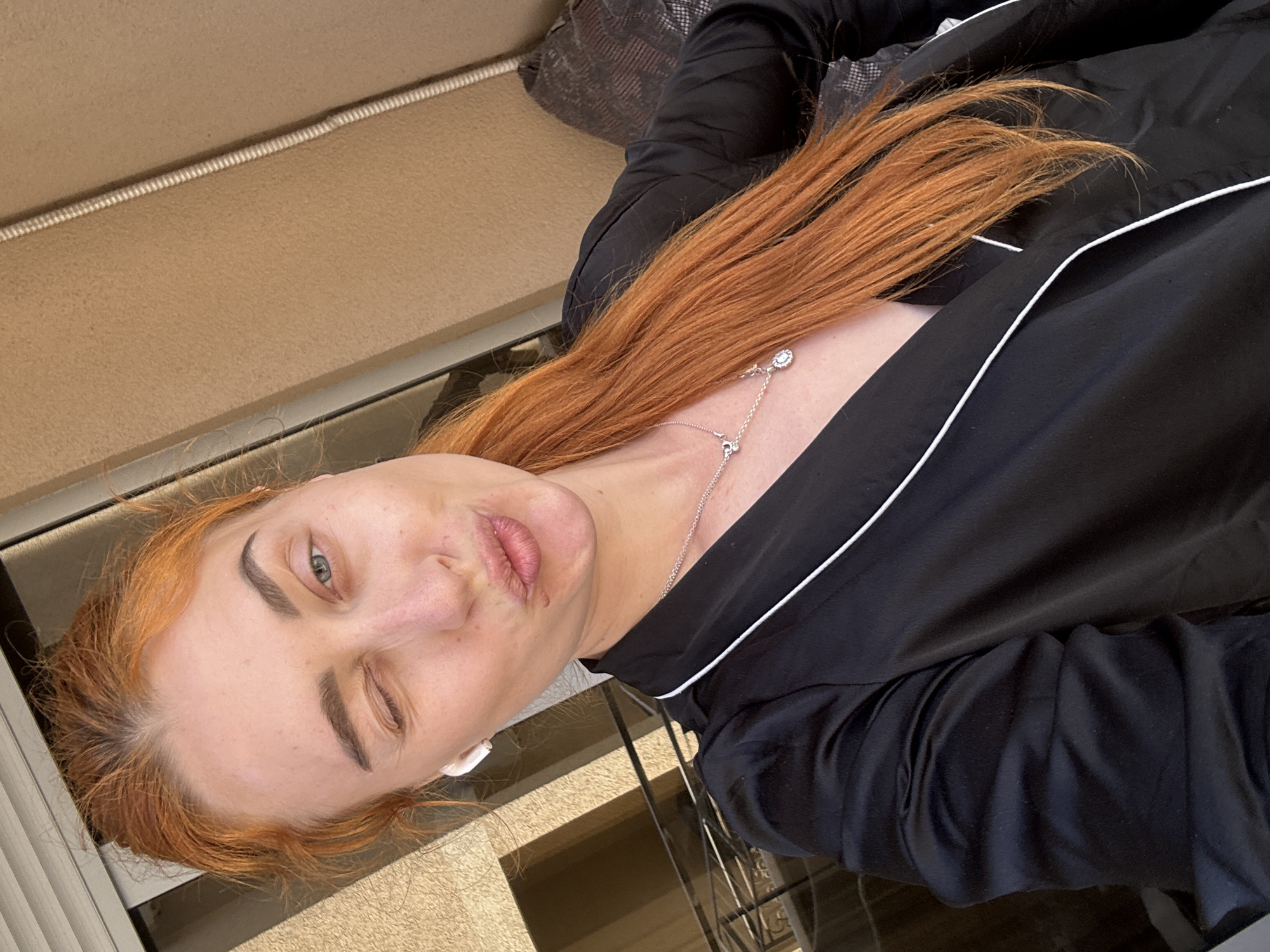A woman with red hair is lying on a tan upholstered surface, winking with her left eye and pouting. She is wearing a black garment with white trim and a silver necklace. An earbud is visible in her left ear. The background includes a patterned pillow and a window or doorway. The scene is likely outdoors. The precise location in Budva, Montenegro, cannot be determined from the image alone.