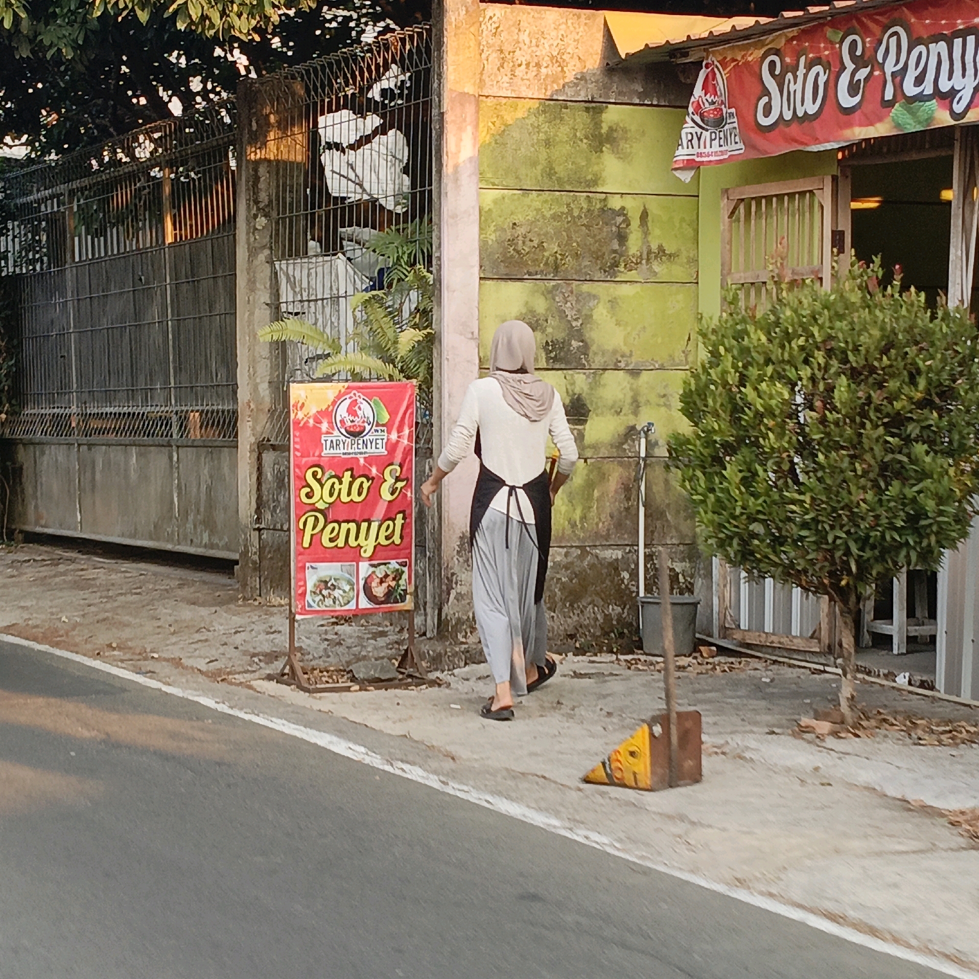 The image shows a woman wearing a hijab, a light-colored long-sleeved shirt, a black apron, and gray pants, walking away from the camera on a sidewalk. She is wearing black flip-flops. In front of her is a red sign with yellow lettering that reads "Soto & Penyett" and displays images of food. To the left of the sign is a metal fence. Behind the woman is a building with a green and yellow facade and a red banner that also says "Soto & Penyett". To the right of the building is a tree. A broom and a yellow wedge are on the sidewalk near the woman. The scene appears to be in an outdoor commercial area. The presence of "Soto & Penyett" signage suggests this is likely a food stall or restaurant. The overall aesthetic and the style of the building are consistent with many establishments found in Salatiga, Indonesia.