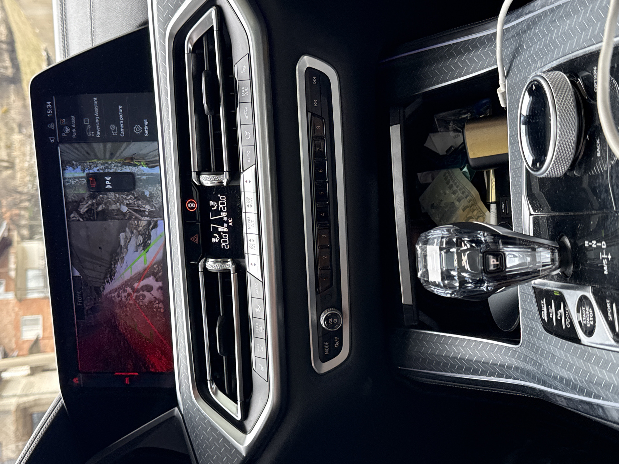 The image shows the interior of a car, most likely a BMW based on the design of the dashboard, infotainment system, and gear selector. The infotainment screen is displaying a camera view labeled "Front," with menu options visible, including "Park Assist," "Reversing Assistant," "Camera picture," and "Settings." The climate control panel indicates a temperature of 20.0°C (68°F). The gear selector is a crystal-style knob with a "P" (Park) indicator. The center console includes buttons for features like "Auto Start/Stop," parking sensors, and a "Sport" mode. The interior trim features a patterned design. The image does not show the location in the city of Lilkovo, Bulgaria.