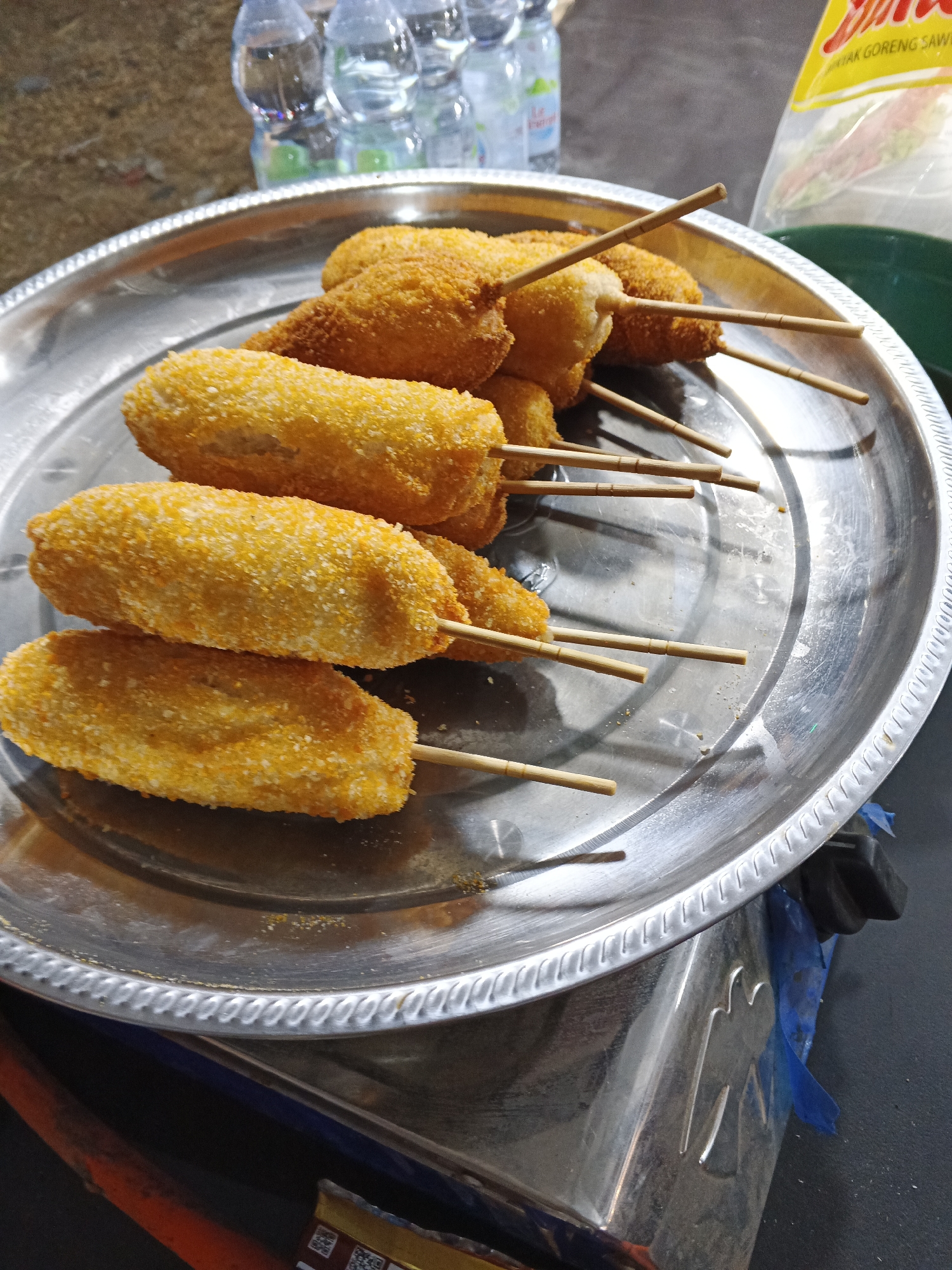 The image displays a platter of golden-brown, breaded food items on skewers, resembling corn dogs, arranged on a silver metallic plate. The food items are cylindrical in shape with a textured, crumb-coated exterior. Several wooden skewers are visible, protruding from the food items. The plate is placed on a dark, textured surface, possibly a table or counter. In the background, several plastic water bottles are visible, along with a yellow plastic bag with Indonesian text that appears to be a food product label. A hint of green liquid is also visible within the bag. The overall setting suggests a casual food stall or eatery.
The scene is indicative of a food vendor in Sukabumi, Indonesia. The presence of Indonesian text on the food packaging and the common street food items like corn dogs are characteristic of such a location. The lighting suggests an outdoor or semi-outdoor setting, possibly at dusk or night, given the reflections on the metallic surfaces and the overall ambient light.