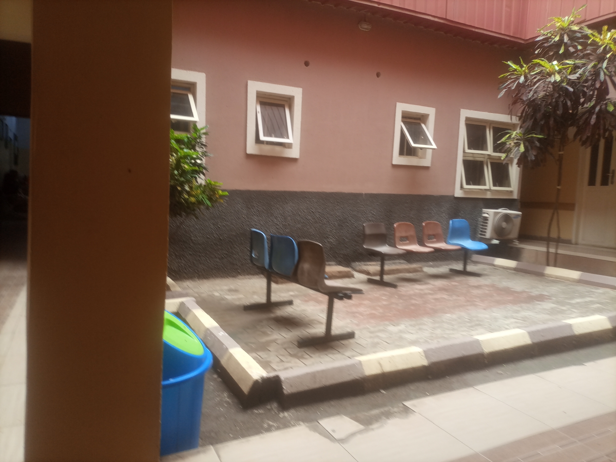 The image depicts an outdoor waiting area in front of a building with a reddish-brown facade and several windows. The ground is paved with interlocking bricks. Several empty chairs, some blue and some brown, are arranged in rows. A blue trash bin with a green lid is visible on the left. An air conditioning unit is mounted on the wall to the right of the chairs. A tree with dark leaves is growing to the right of the building. The scene appears to be located in the city of Tumfure, Nigeria.